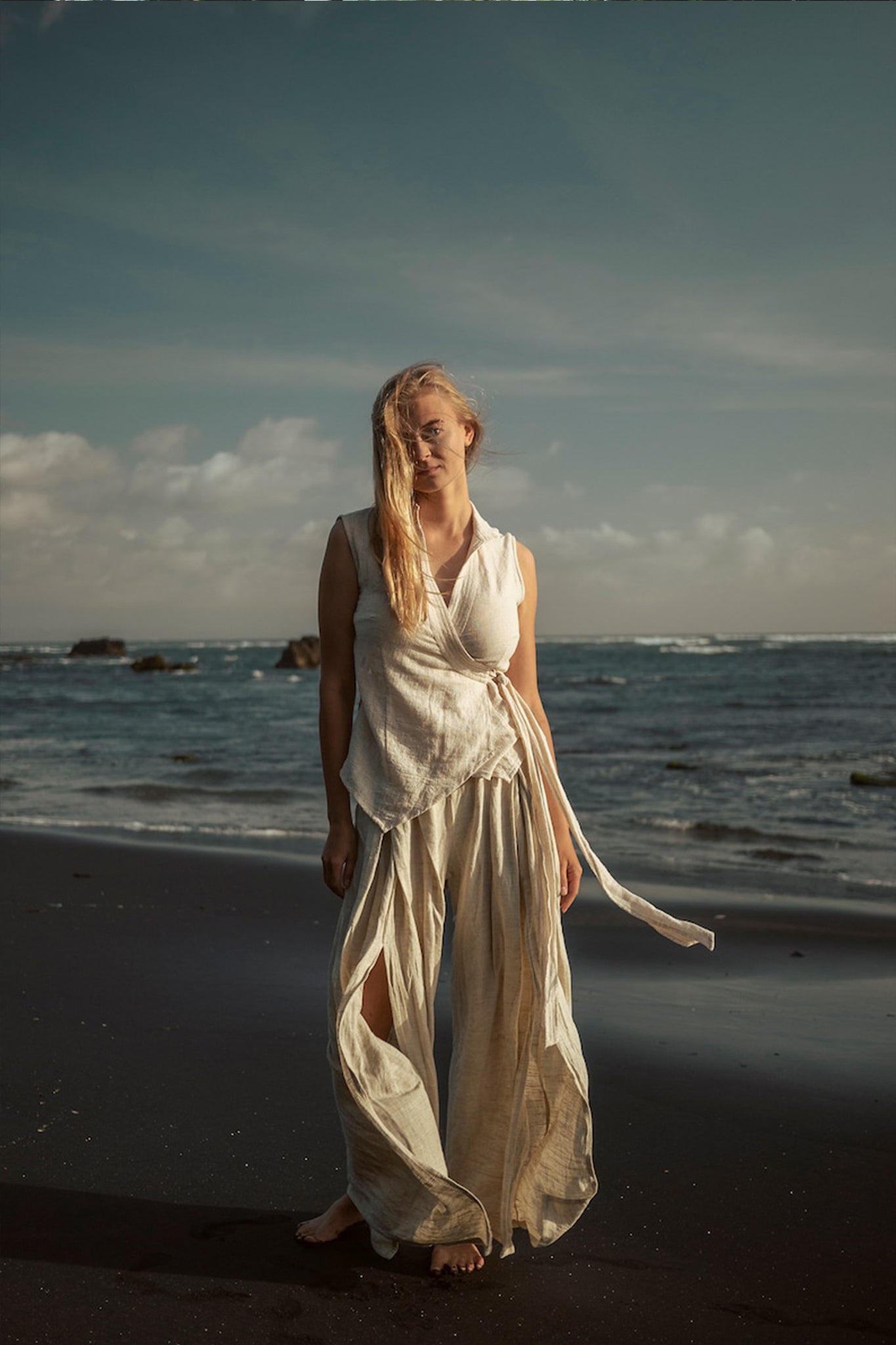 A person standing barefoot on a beach, dressed in a light, flowing white top paired with AYA Sacred Wear's Off-White Linen Boho Pants with High Slit. The ocean waves gently crashing under a partly cloudy sky create a serene and peaceful atmosphere that perfectly embodies sustainable fashion.