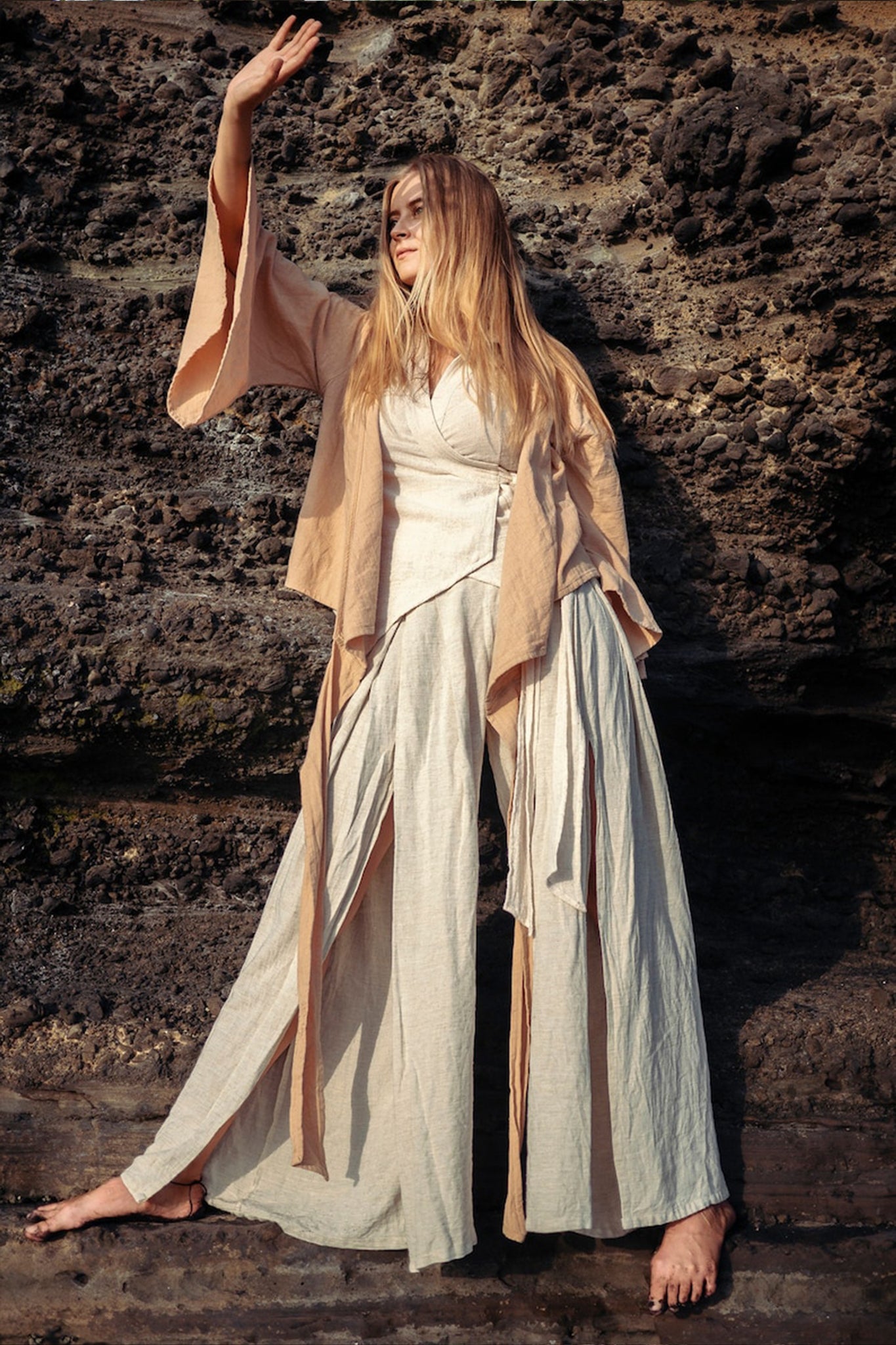 Standing against a rugged stone wall, a person with long hair showcases the essence of sustainable fashion by wearing AYA Sacred Wear's Off-White Linen Boho Pants with High Slit. With one arm raised, the natural texture of the rock provides a striking contrast to the airy elegance of these wide-legged trousers.