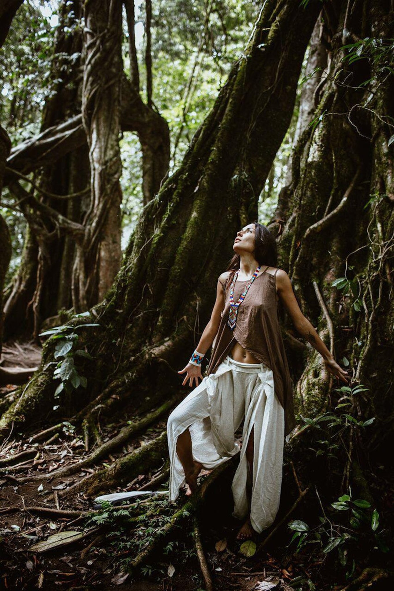 A person stands in a lush forest, surrounded by large tree roots and dense foliage. They are wearing a brown sleeveless top paired with AYA Sacred Wear's Off-White Linen Boho Pants with High Slit, looking upwards with arms outstretched. Sunlight filters through the trees, casting a warm glow on their embrace of sustainable fashion.