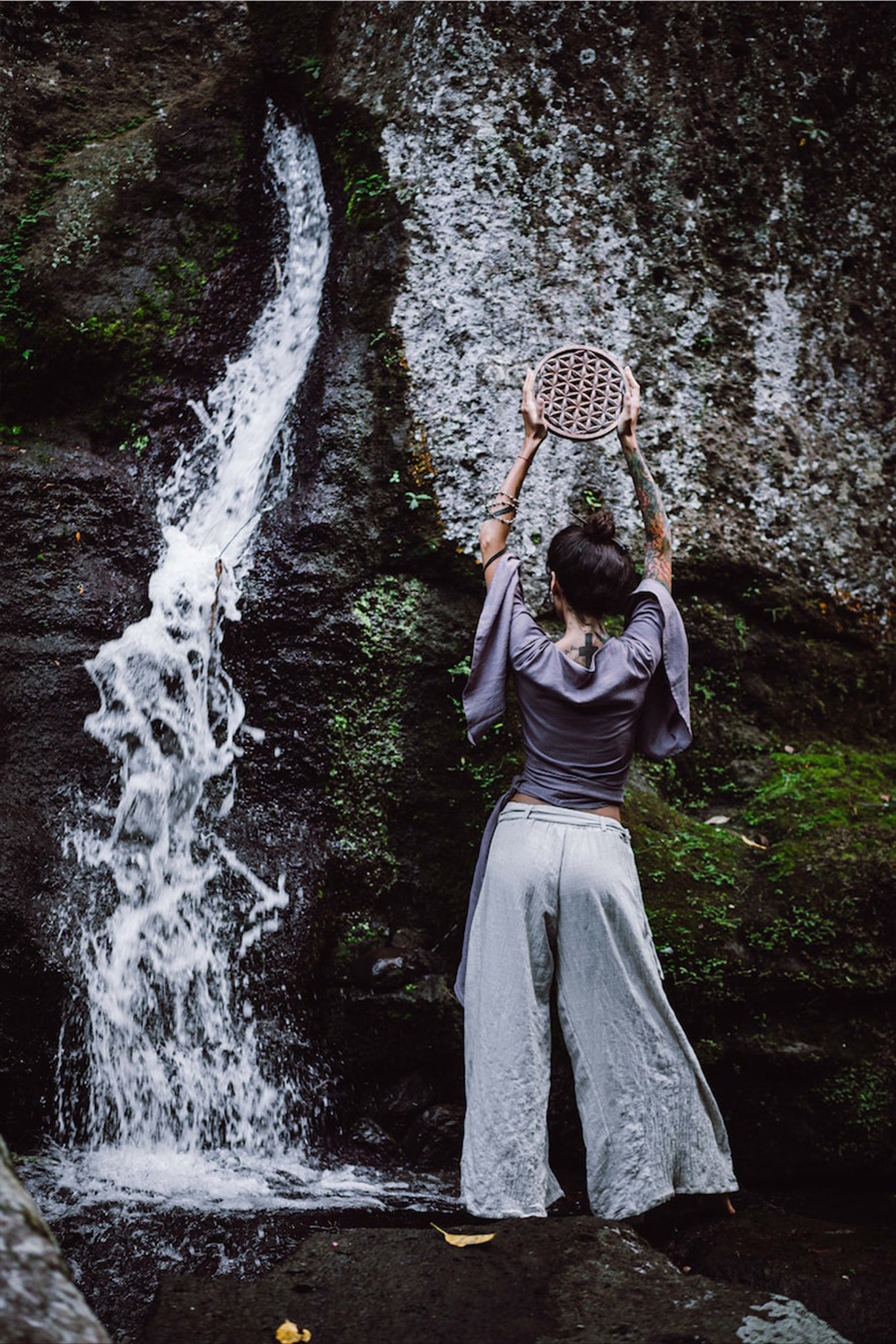 A tattooed individual stands facing a rocky waterfall, holding a circular wooden object above their head. They are wearing AYA Sacred Wear's Off-White Linen Boho Pants with High Slit and a dark top, surrounded by lush greenery, embodying the essence of sustainable fashion.
