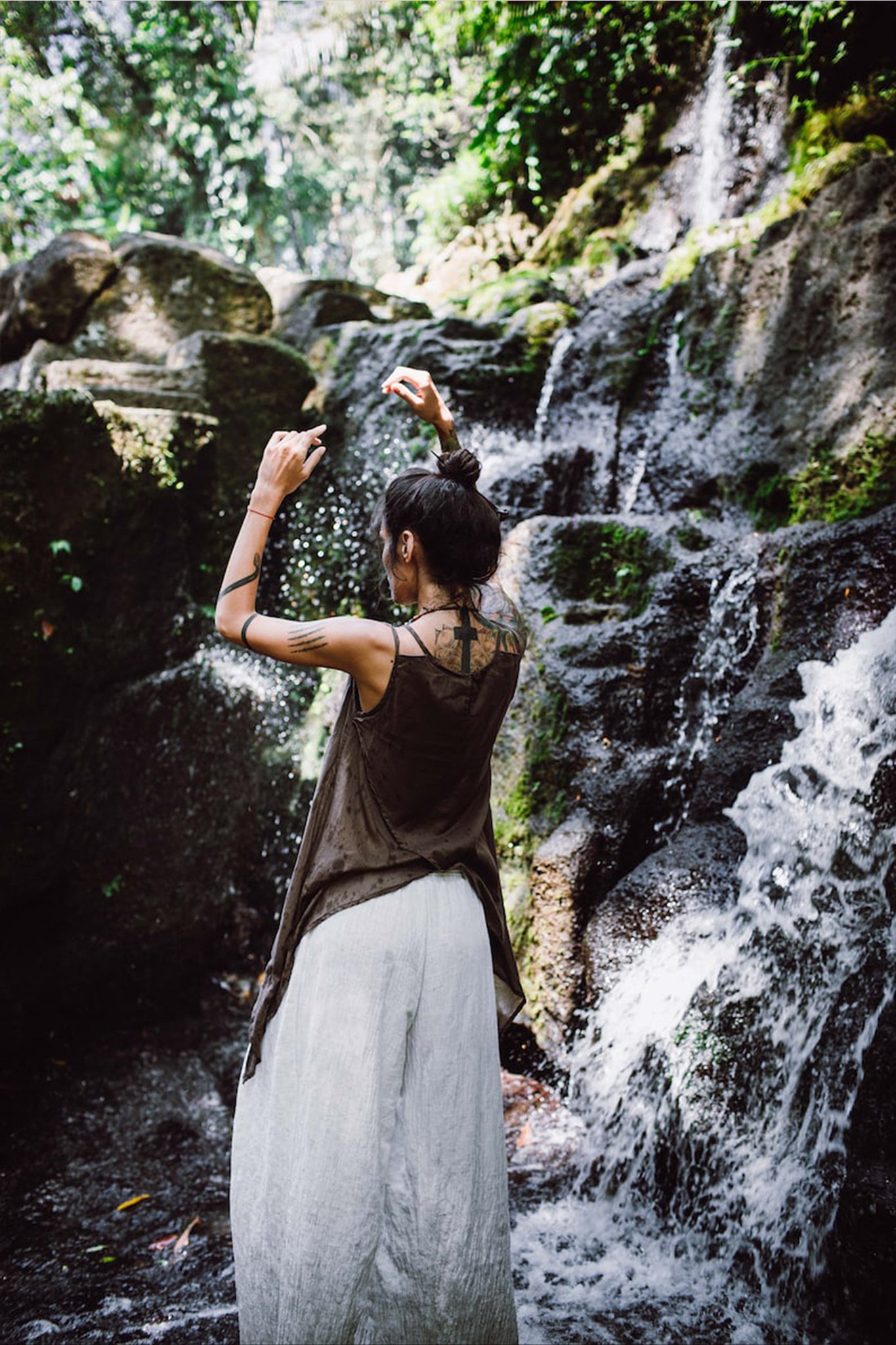 Clad in the Off-White Linen Boho Pants with High Slit from AYA Sacred Wear, an individual with long hair tied up stands in front of a moss-covered rocky waterfall. Their arms are raised as sunlight filters through the surrounding greenery, illuminating the cascading water.