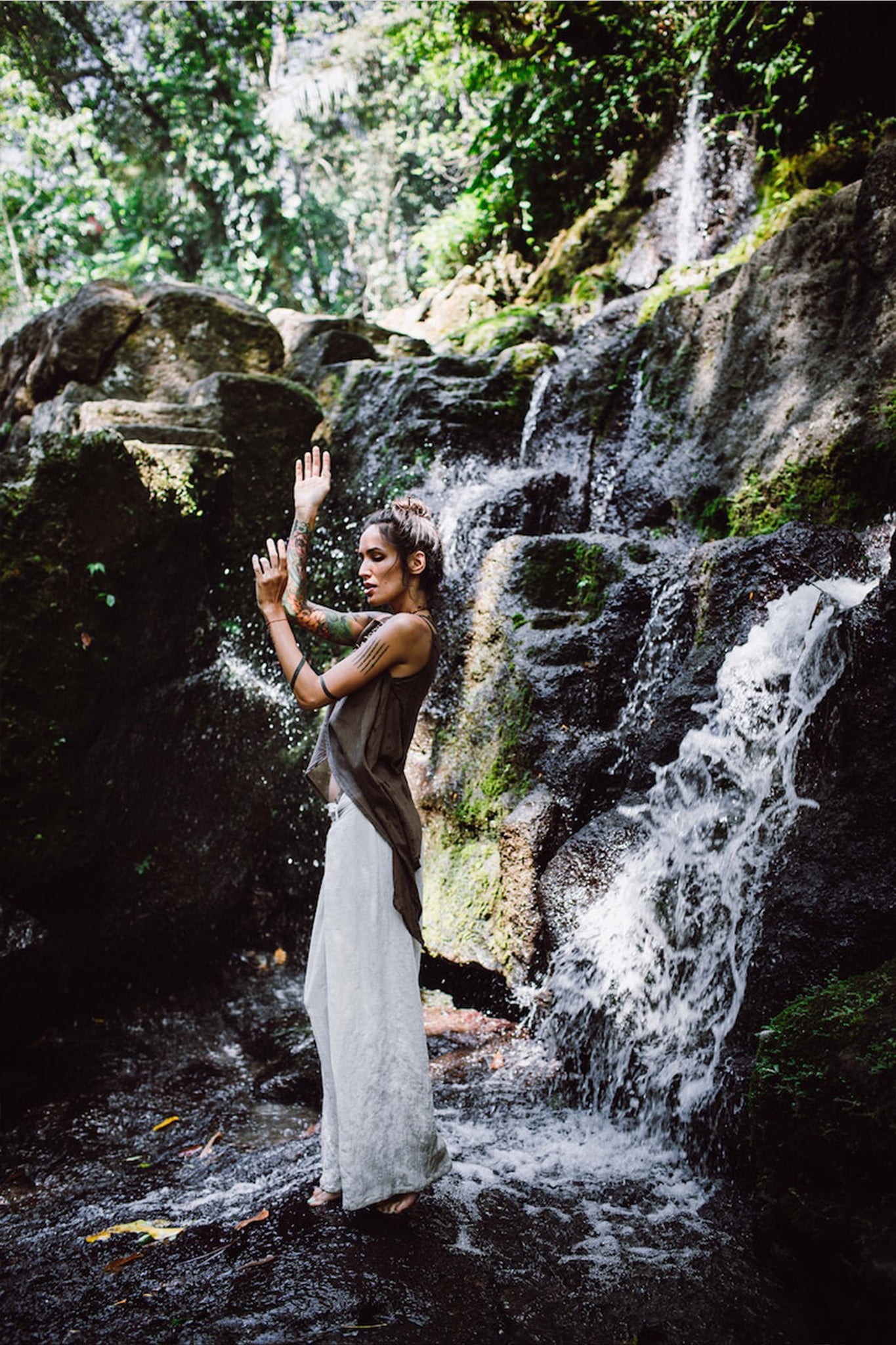 Amidst a lush, green forest beside a small waterfall, a woman poses elegantly in off-white linen boho pants with a high slit by AYA Sacred Wear. The sunlight filtering through the trees highlights her commitment to sustainable fashion as it illuminates the water and surrounding foliage.