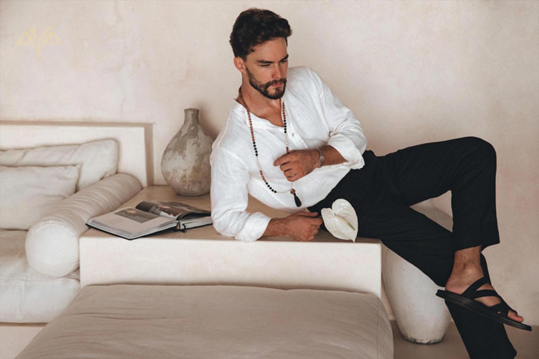 A man in an Off-White Linen Classic Minimalist Shirt from AYA Sacred Wear is lounging on a minimalist, beige sofa, holding a hat. An open book and a decorative vase are beside him, creating a calm and relaxed setting.