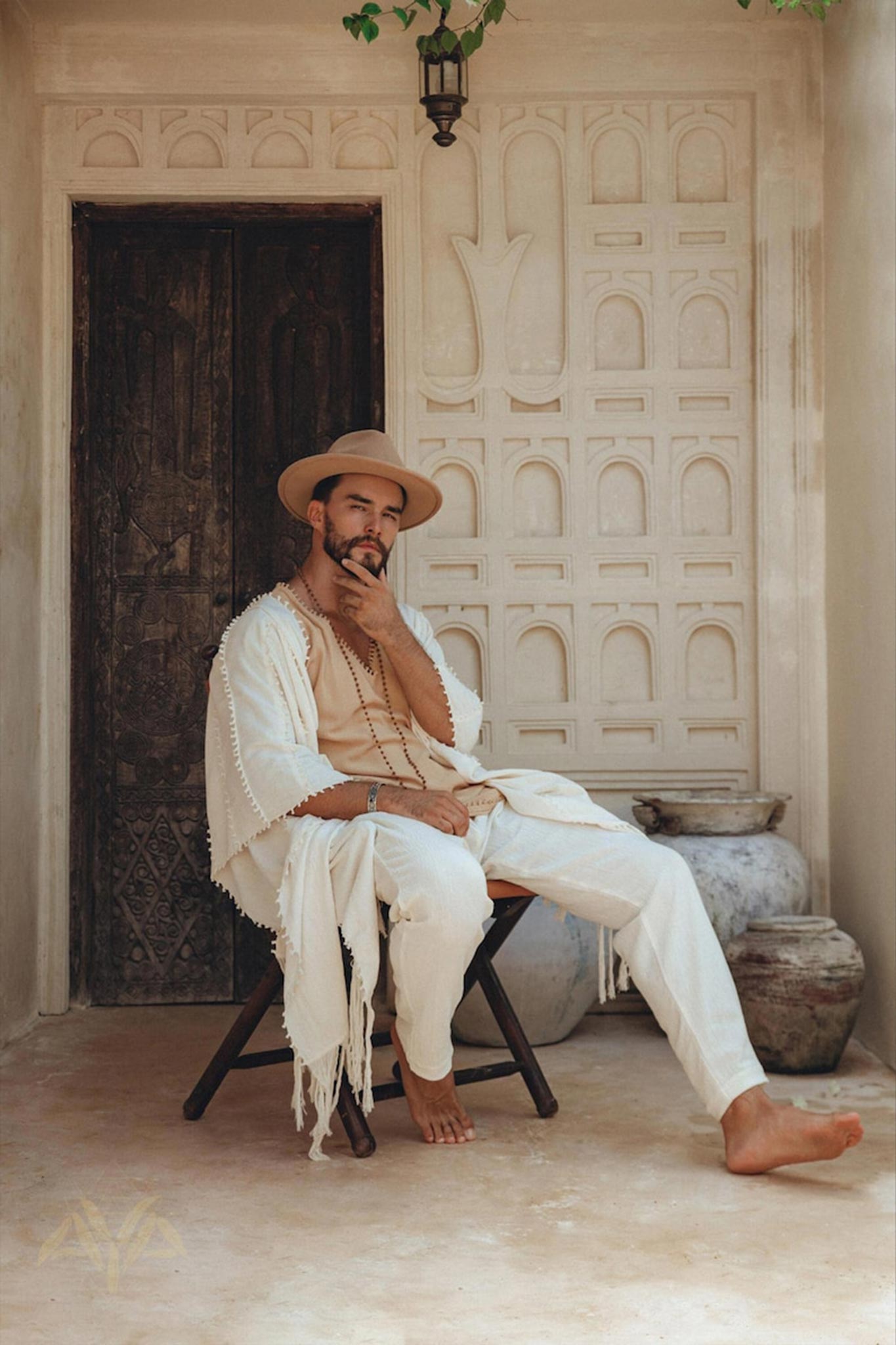 A man in a beige outfit and hat sits on a wooden chair in front of an ornate door. His ensemble includes AYA Sacred Wear's Off-White Long Cotton Pants for Men, blending seamlessly with earthy tones, clay pots, and intricate wall designs, creating a serene, rustic atmosphere.