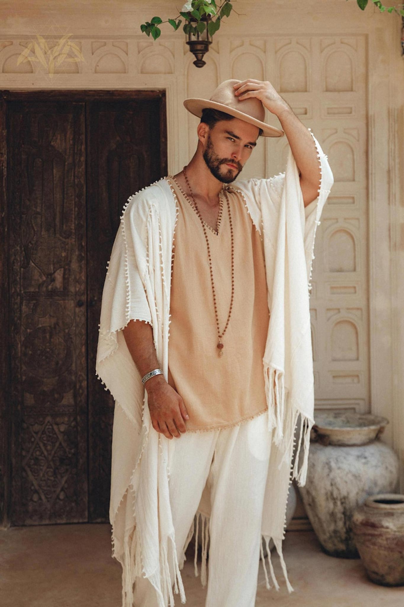 A man stands in front of an ornate wooden door wearing a beige shirt, a botanically dyed shawl, and a hat. His left hand rests on the brim of his hat as he accessorizes with a beaded necklace and Off-White Long Cotton Pants by AYA Sacred Wear. Two large clay pots are visible in the background.