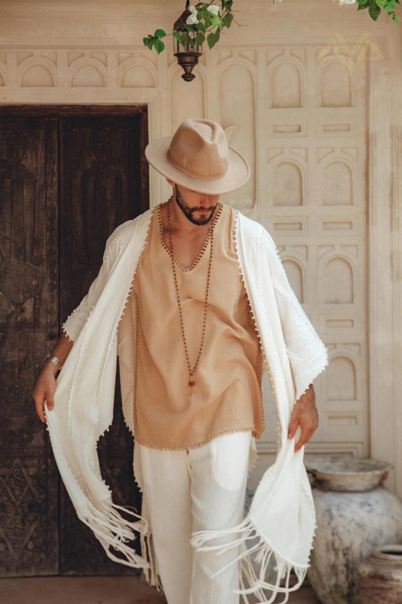 A man clad in an AYA Sacred Wear tan hat, beige shirt, Off-White Long Cotton Pants for Men, long necklace, and white fringed shawl stands before an intricately designed door. Gazing downward with one hand holding the shawl open, he is framed by a hanging lantern and potted plant that enhance the serene scene.