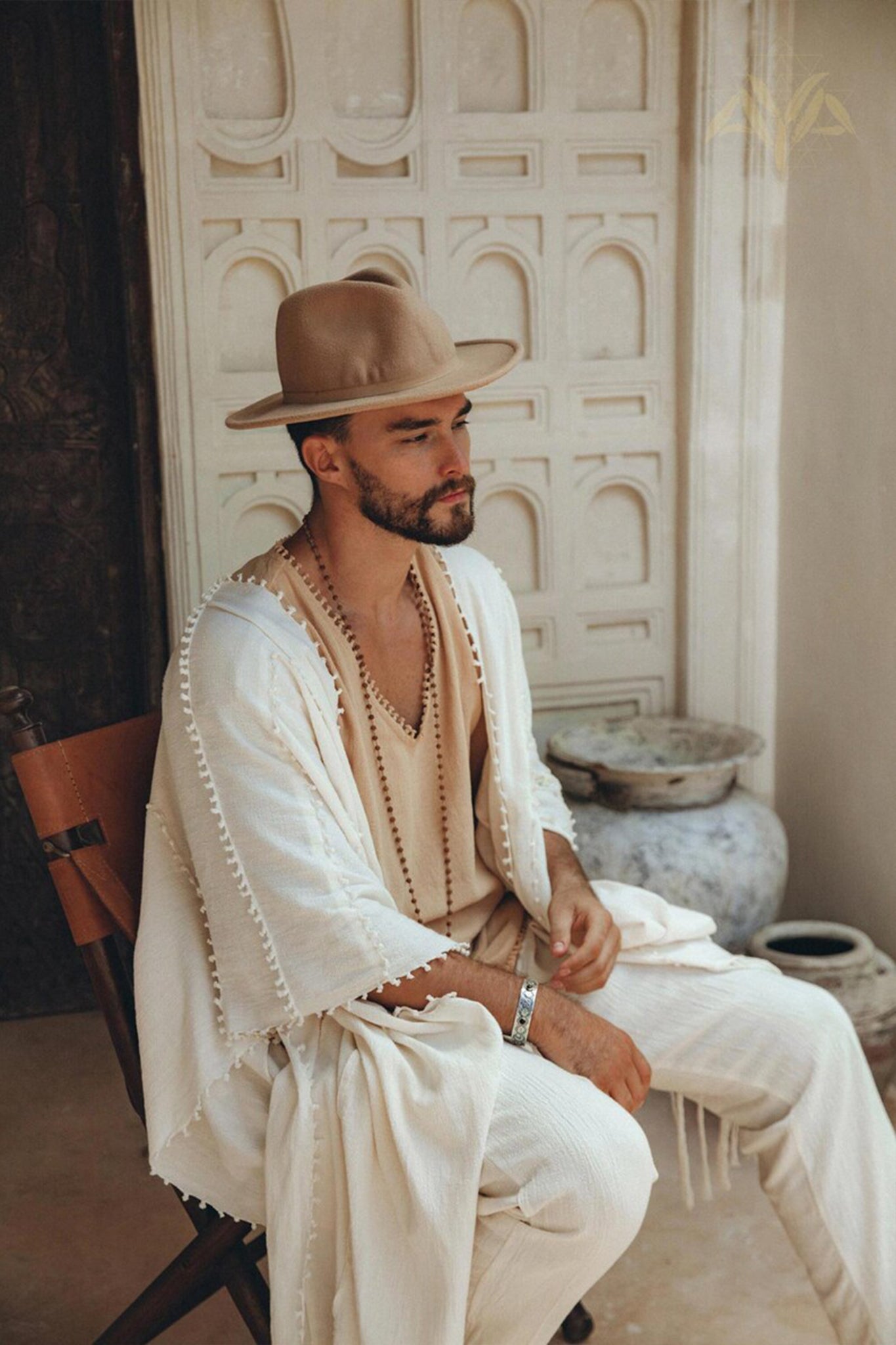 A bearded man dressed in neutral tones, including a wide-brimmed hat and a robe over AYA Sacred Wear's Off-White Long Cotton Pants for Men, sits on a wooden chair. The scene is set against an ornate white wall with architectural details, creating a serene and stylish ambiance.