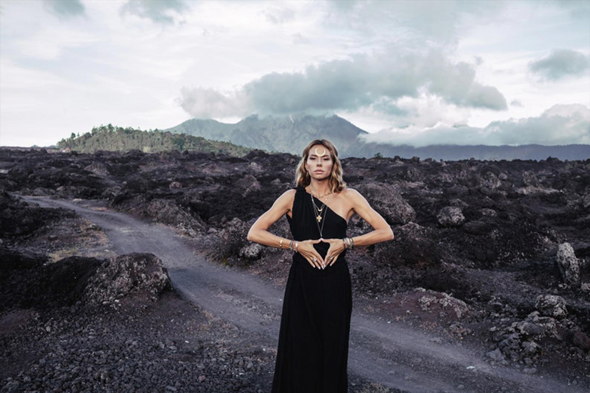 A person in an Organic Black Boho One Shoulder Dress by AYA Sacred Wear stands on a rocky landscape with a volcanic mountain in the background. They form a heart shape with their hands at their waist, highlighted by a braided belt. The sky above is cloudy.