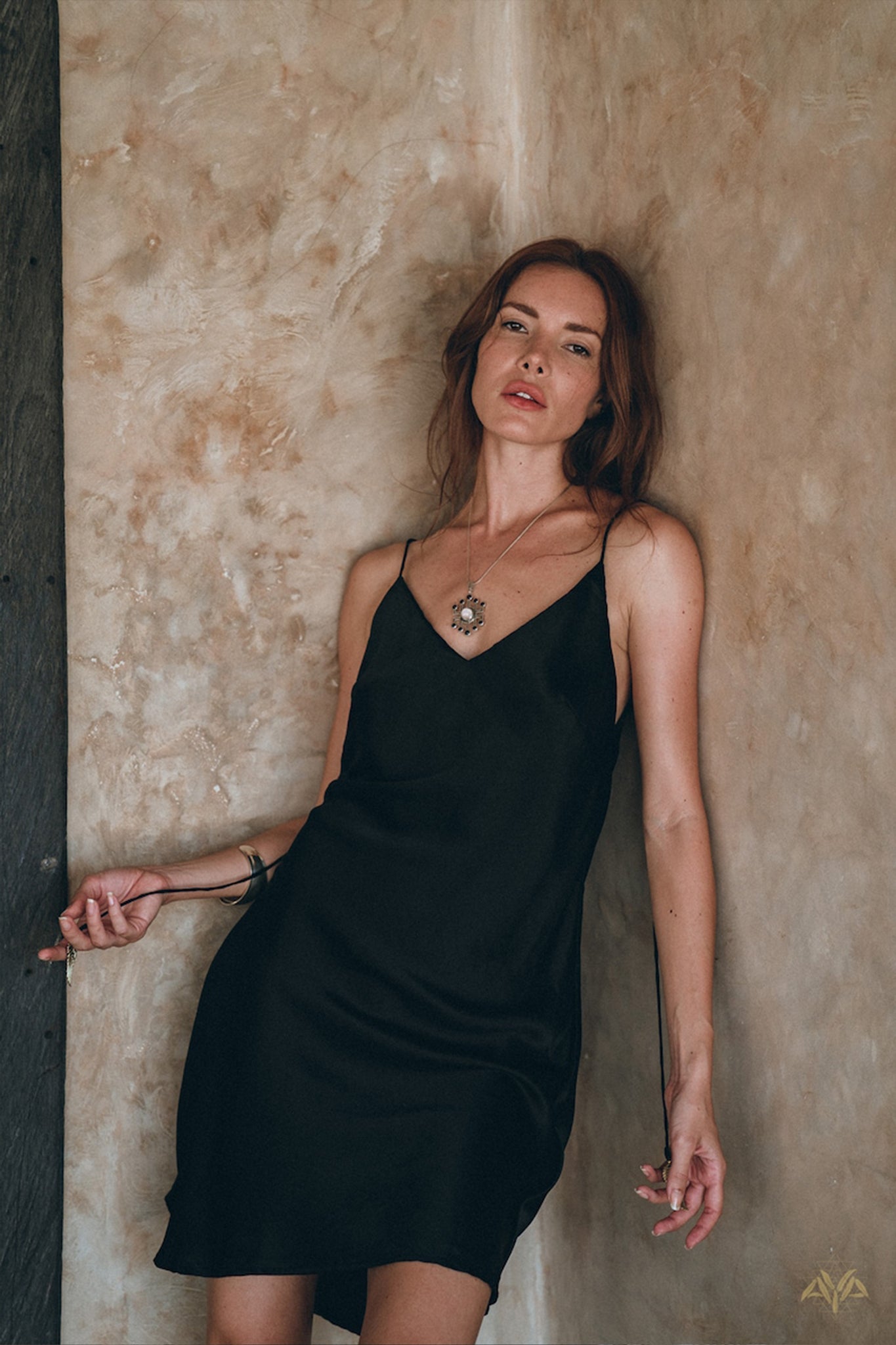 A woman wearing an AYA Sacred Wear Peace Silk Slip Dress in pure organic silk leans against a beige textured wall, her left hand resting on her hip and her right hand touching the wall. She accessorizes with a silver necklace and has long, wavy brown hair.