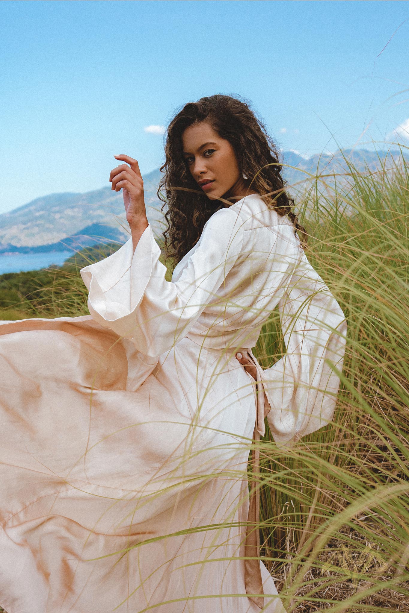 A model poses gracefully in a field, wearing the Powder Pink Venus Peace Silk Skirt, a minimalist wrap skirt that flows elegantly with her movements. The skirt's organic fabric complements the natural landscape, featuring lush grass and a scenic backdrop. The soft pastel hue and stylish design make it a perfect choice for a summer outfit.