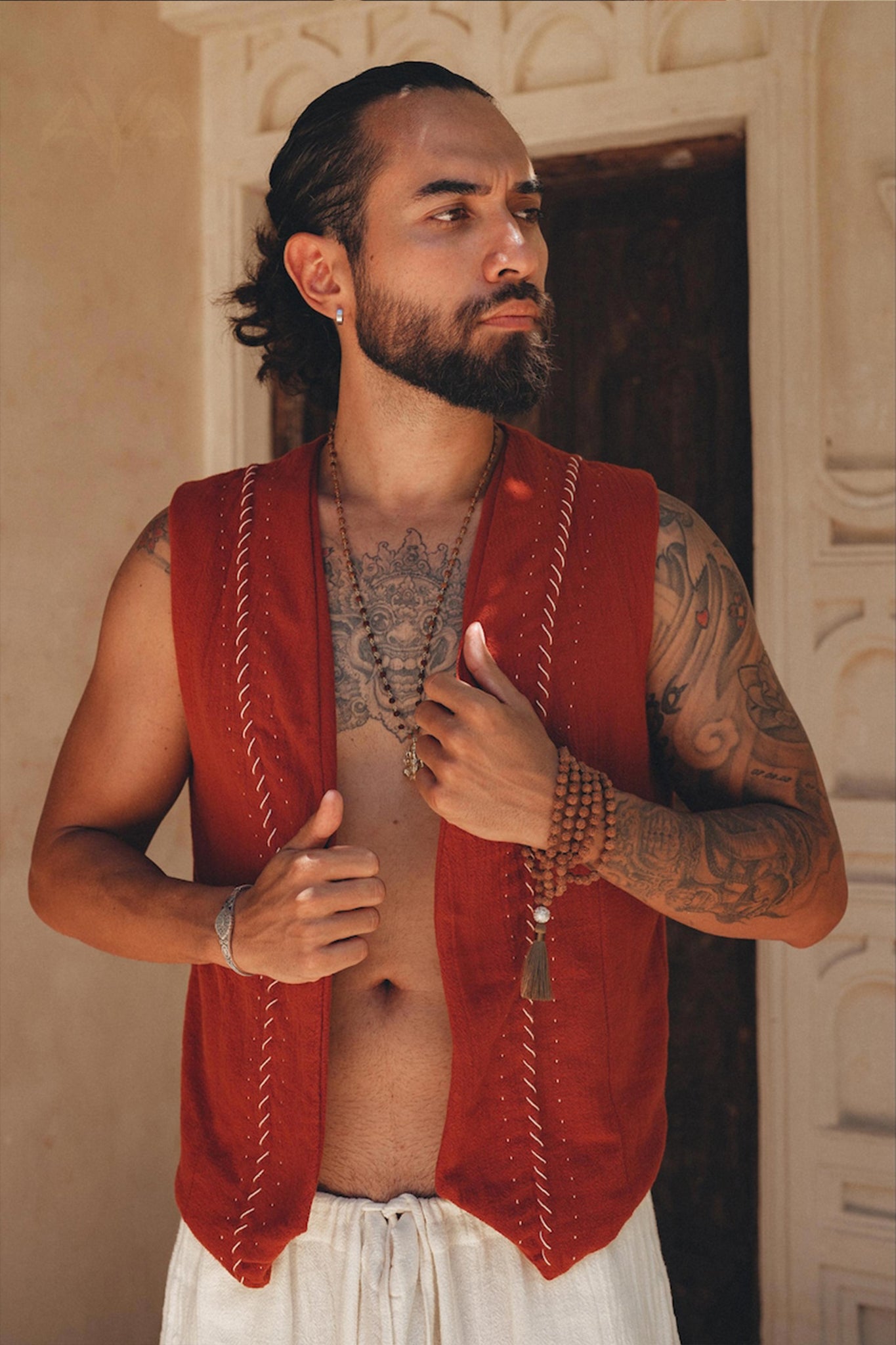 A person with long, dark hair tied back is wearing a Red Hand Embroidered Vest for Men from AYA Sacred Wear and white pants, standing in front of an ornate wall. They are adorned with tattoos on their arms and chest and are holding a beaded bracelet in one hand.