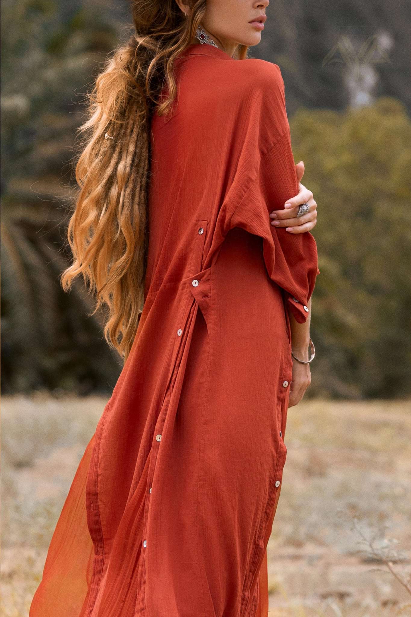 In a serene outdoor setting, an individual with long, wavy hair wears AYA Sacred Wear's Red Kannika Shirt Dress, made from lightweight fabric and botanical dyes. The dress features a multiway buttoned back design against blurred greenery.
