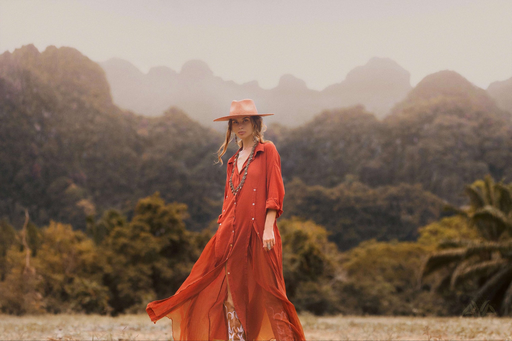 In a misty field with mountains behind, a person dons an AYA Sacred Wear Red Kannika Shirt Dress—an adjustable, multiway button-down crafted from organic cotton and enhanced by botanical dyes—paired with a wide-brimmed hat, imparting serene elegance.