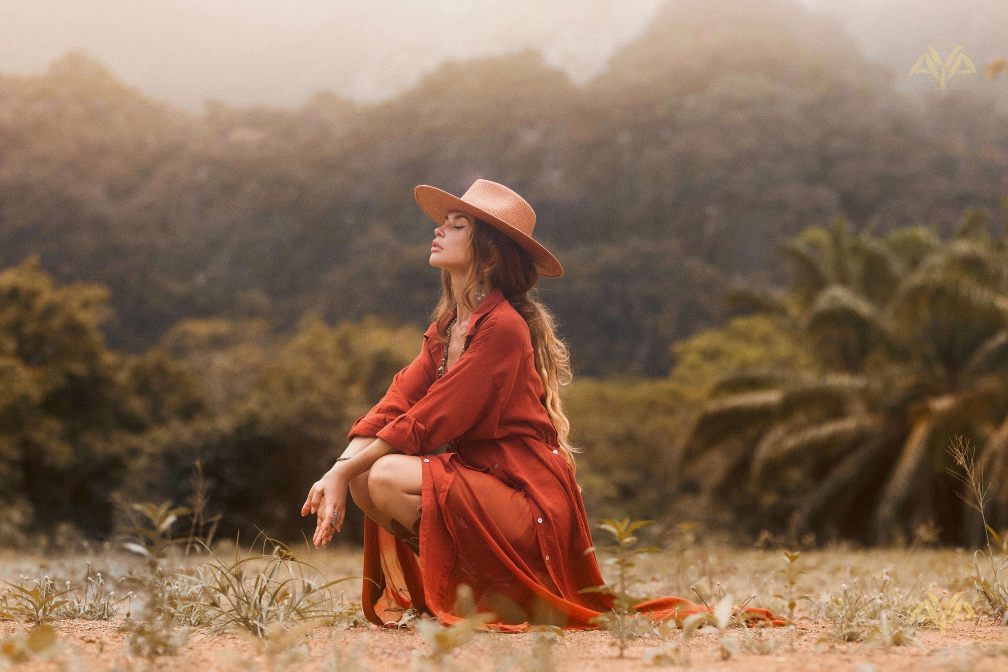 Kneeling amidst nature, a woman in AYA Sacred Wear's Red Kannika Shirt Dress, made from organic cotton with botanical dyes, relaxes with eyes closed under a misty sky of trees and mountains.