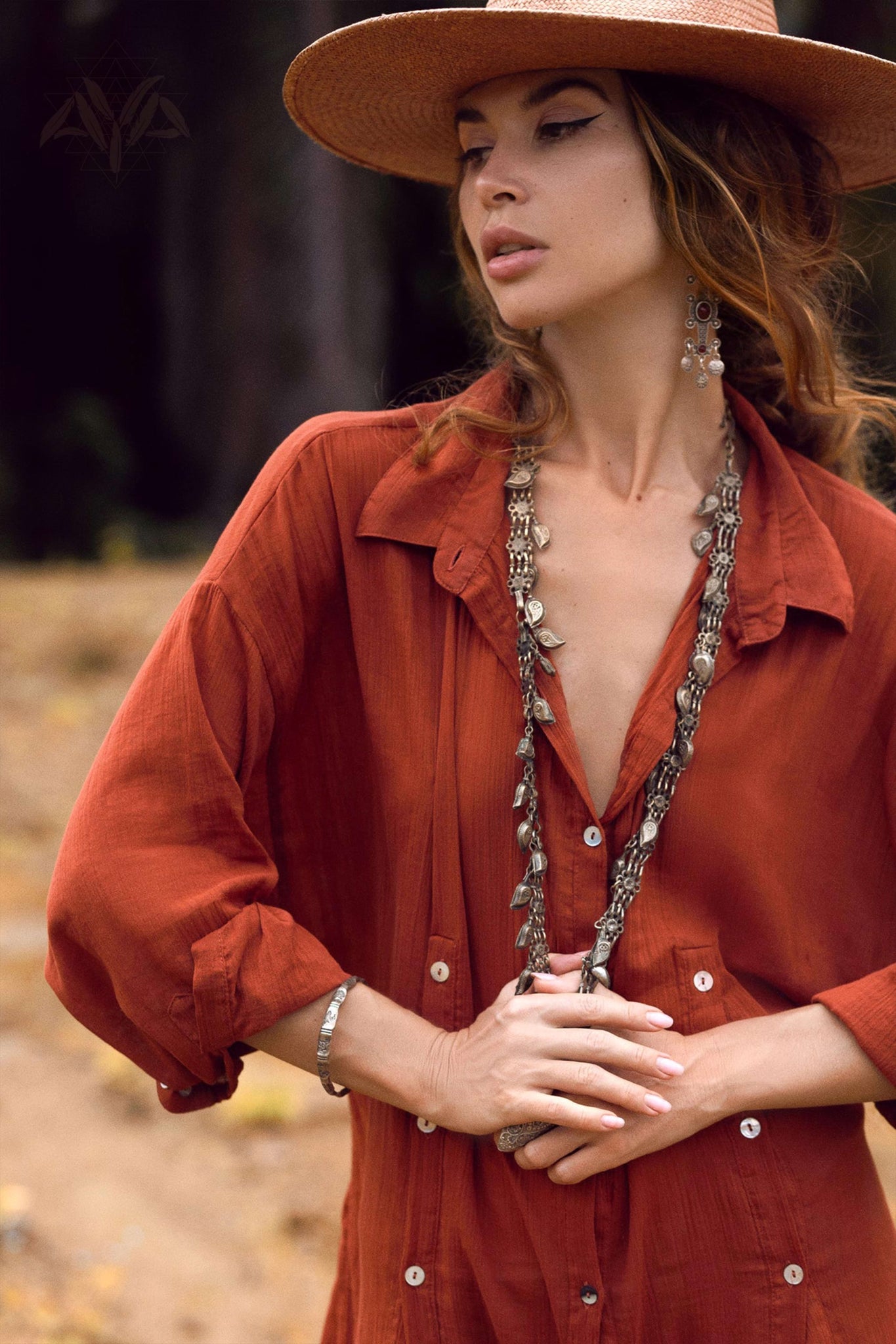 A woman outdoors in an orange button-up dress, similar to AYA Sacred Wear's Red Kannika Shirt Dress, with a wide-brimmed straw hat. She's accessorized with long necklaces, intricate earrings, and a bracelet. Her gaze is directed sideways against a backdrop of blurred foliage.