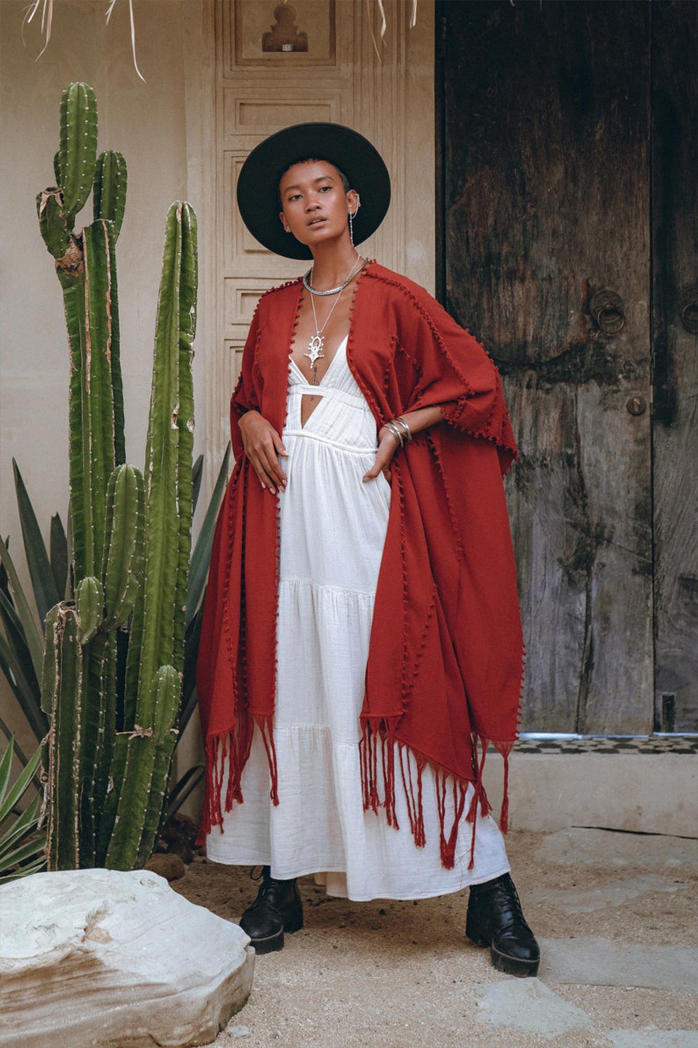 In a rustic setting featuring a wooden door and large cactus, a woman showcases her style with a white dress and red poncho robe from AYA Sacred Wear. She pairs it with a black hat, boots, and silver necklace, creating an ensemble that perfectly complements the desert-like ambiance.
