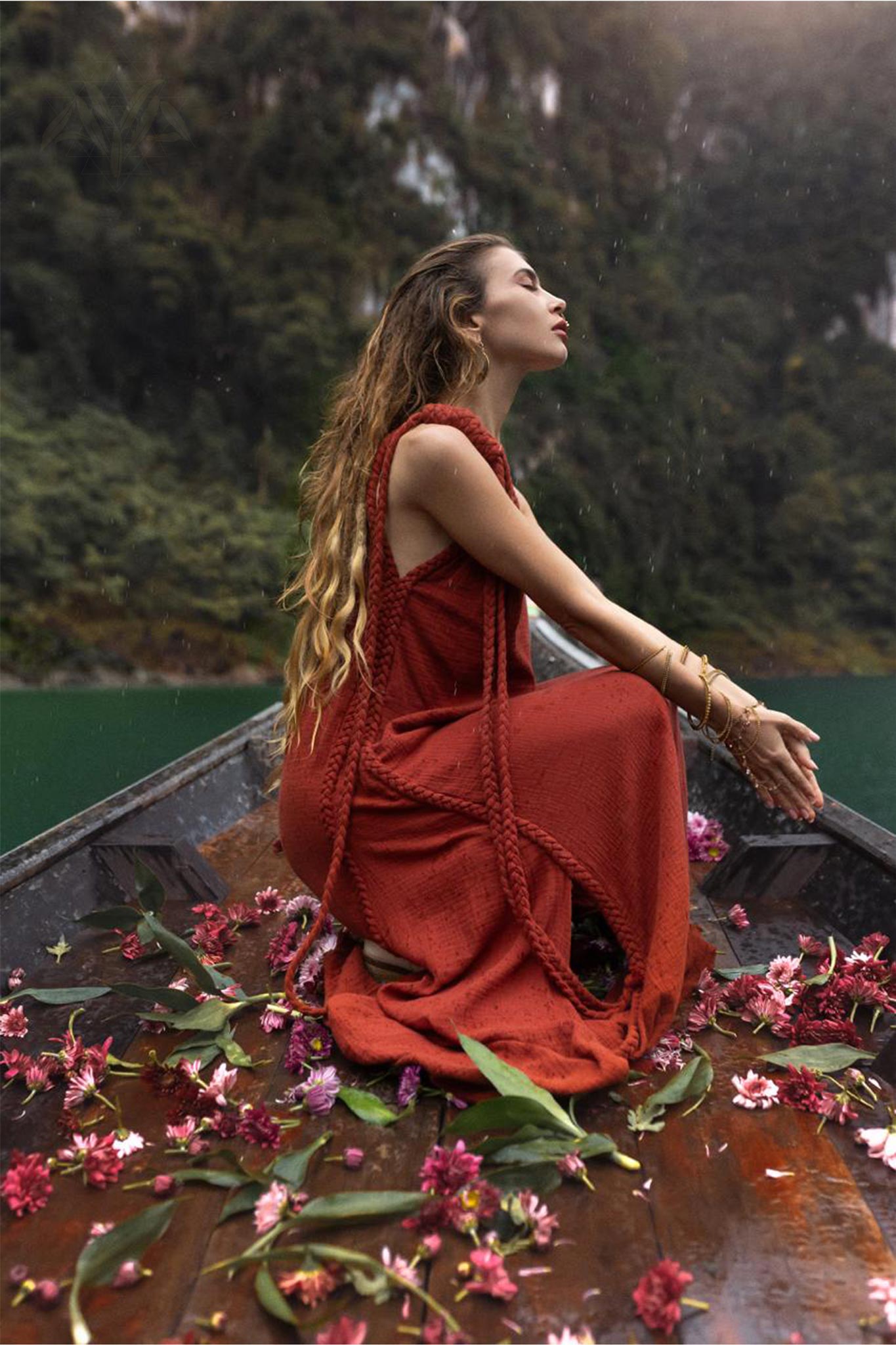 A woman with long, wavy hair wearing an AYA Sacred Wear Red Aurora Greek Goddess Dress • Boho Macrame Dress • Boho Sustainable Dress sits at the end of a wooden boat. She is surrounded by scattered flowers and greenery on the boat's deck. The background features lush green foliage and the calm water of a lake or river.