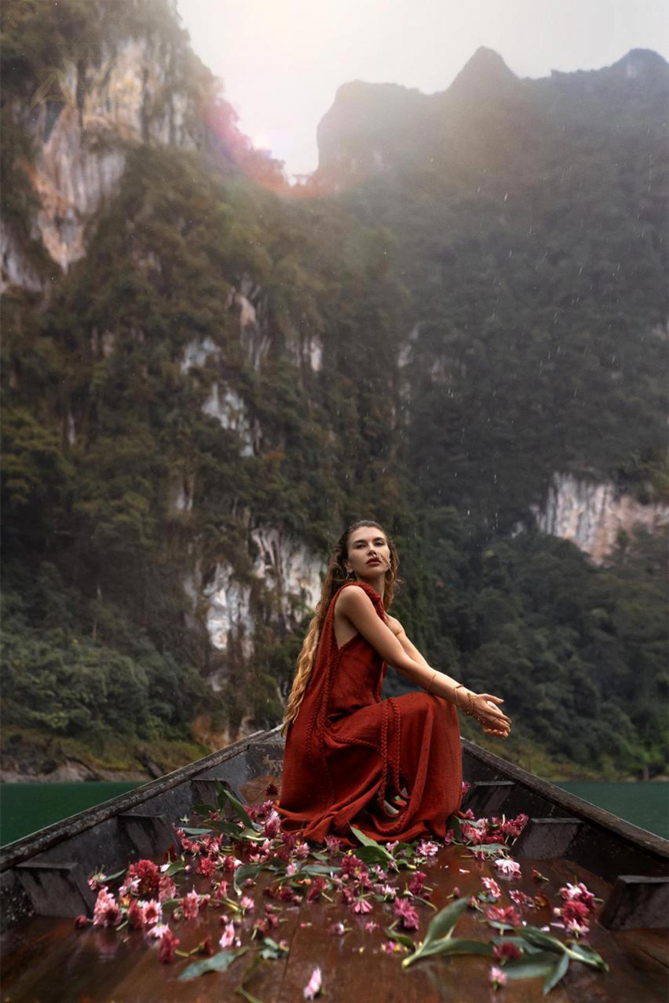 A woman in a flowing Red Aurora Greek Goddess Dress by AYA Sacred Wear, complete with long braids, sits peacefully at the bow of a boat adorned with scattered pink and red flowers. Steep green cliffs rise dramatically behind her under a cloudy sky, creating a serene and mystical atmosphere.