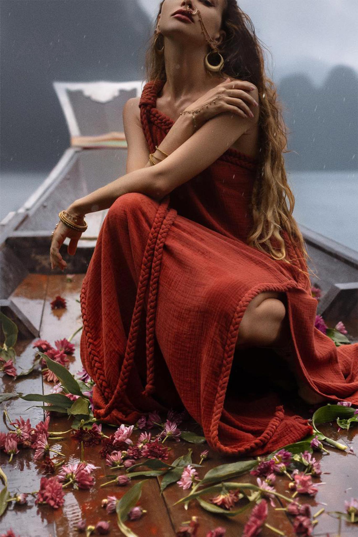 A person wearing the Red Aurora Greek Goddess Dress by AYA Sacred Wear sits on a wooden boat adorned with scattered pink flowers and green leaves. They look upward, crossing their arms gently. The backdrop appears to be a misty, serene lake.