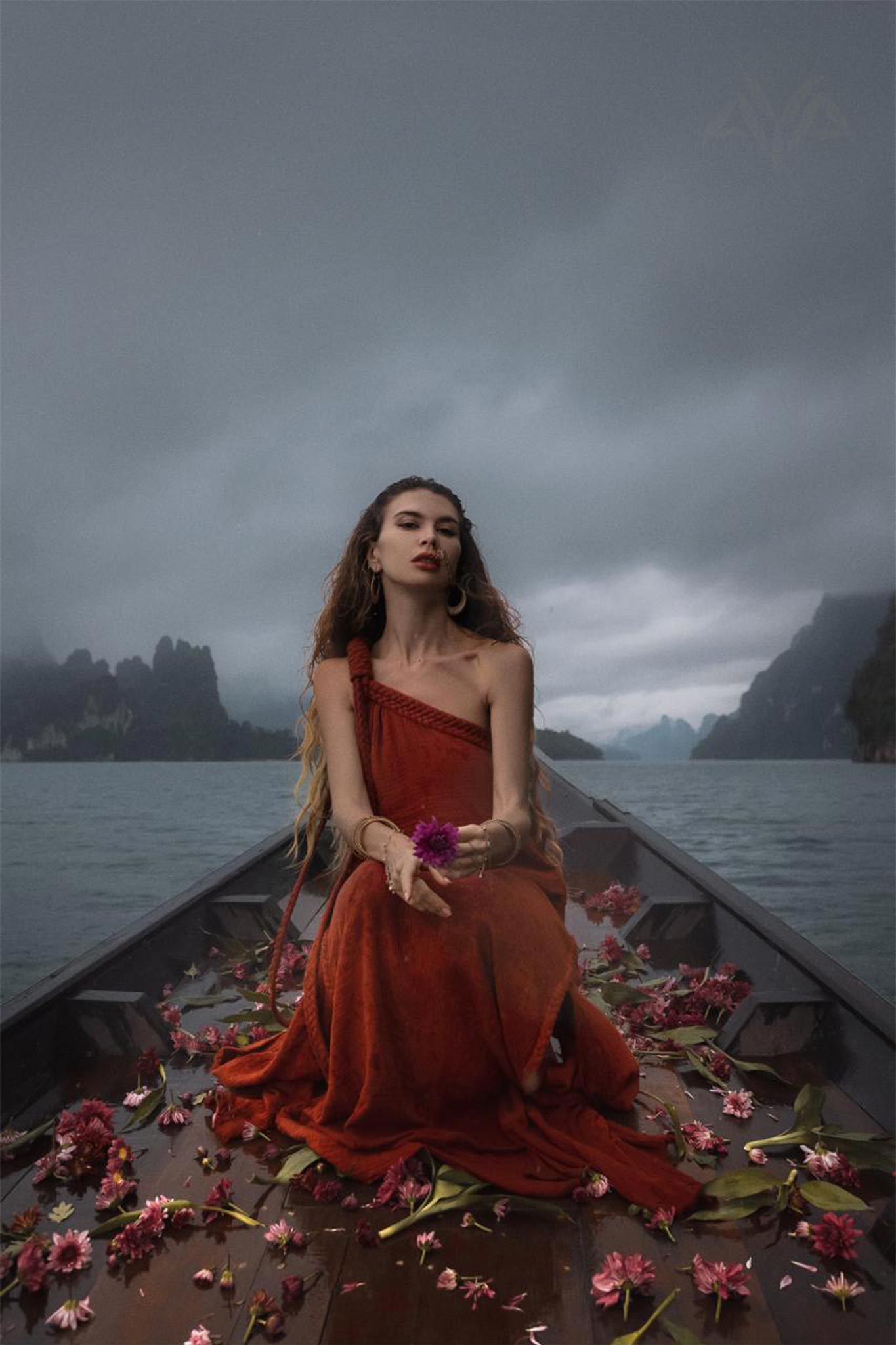 A woman in an AYA Sacred Wear Red Aurora Greek Goddess Dress sits on a boat surrounded by water and misty mountains. She holds a flower, with more scattered around her. The atmosphere is serene and mystical, with overcast skies adding a moody effect to the scene.