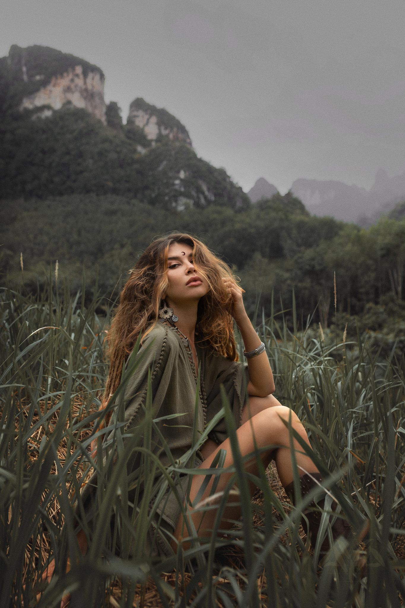A woman with wavy hair in a sage green, hand-knotted romper sits in tall grass with a mountainous landscape in the background.