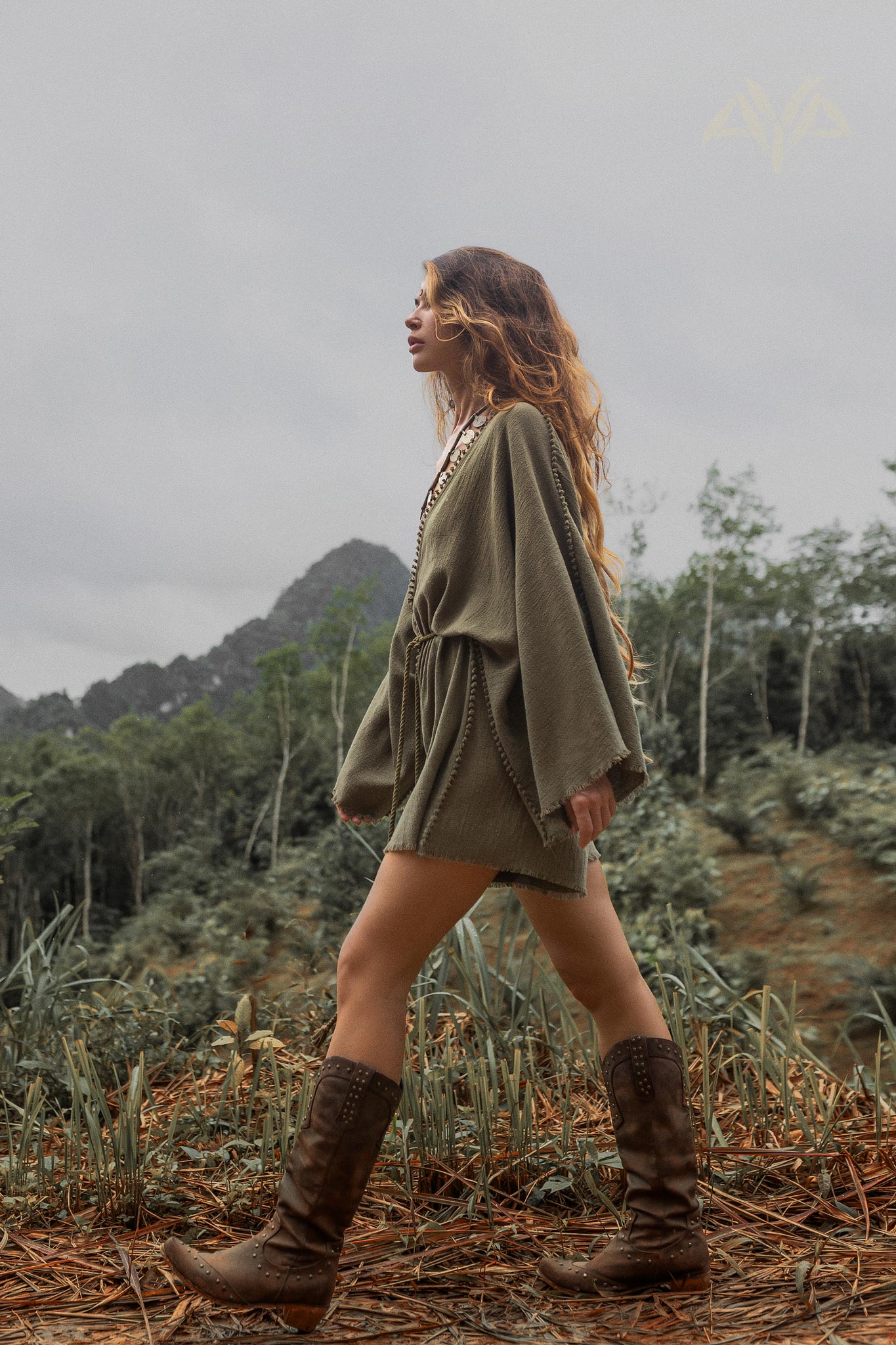 A woman walking through a natural outdoor environment, wearing a sage green hand-knotted mini organic jumpsuit romper dress. The outfit features flowing bell sleeves and a cinched waist. She completes her look with tall brown boots adorned with studs. Her long, wavy hair flows naturally as she strides confidently across the earthy terrain, with a backdrop of green trees and distant hills under a cloudy sky.