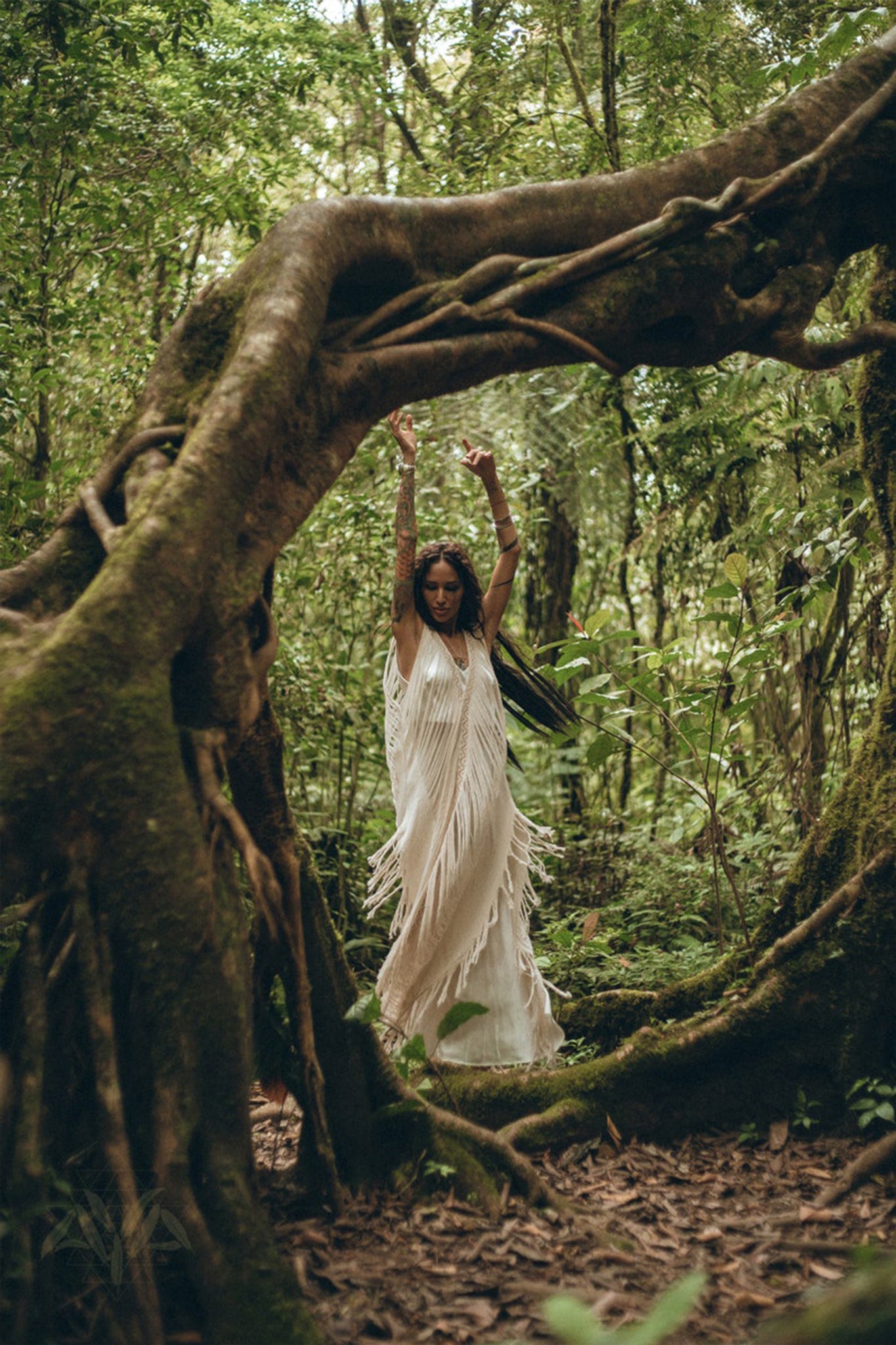A woman in an AYA Sacred Wear See Through Dress, a long maxi dress made of tribal raw cotton, dances gracefully in a lush green forest, surrounded by twisting tree roots and foliage. Her arms are raised, and the scene is serene and mystical.