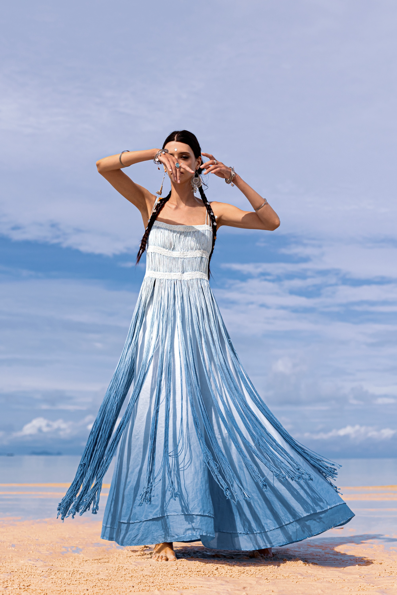 A woman stands barefoot on a sandy beach, wearing an AYA Sacred Wear Sky Blue Greek Goddess Dress botanically dyed in blue shades of organic cotton. Her braided hair and tribal bracelets sway in the gentle breeze, under a sky dotted with clouds as the ocean softly whispers nearby.