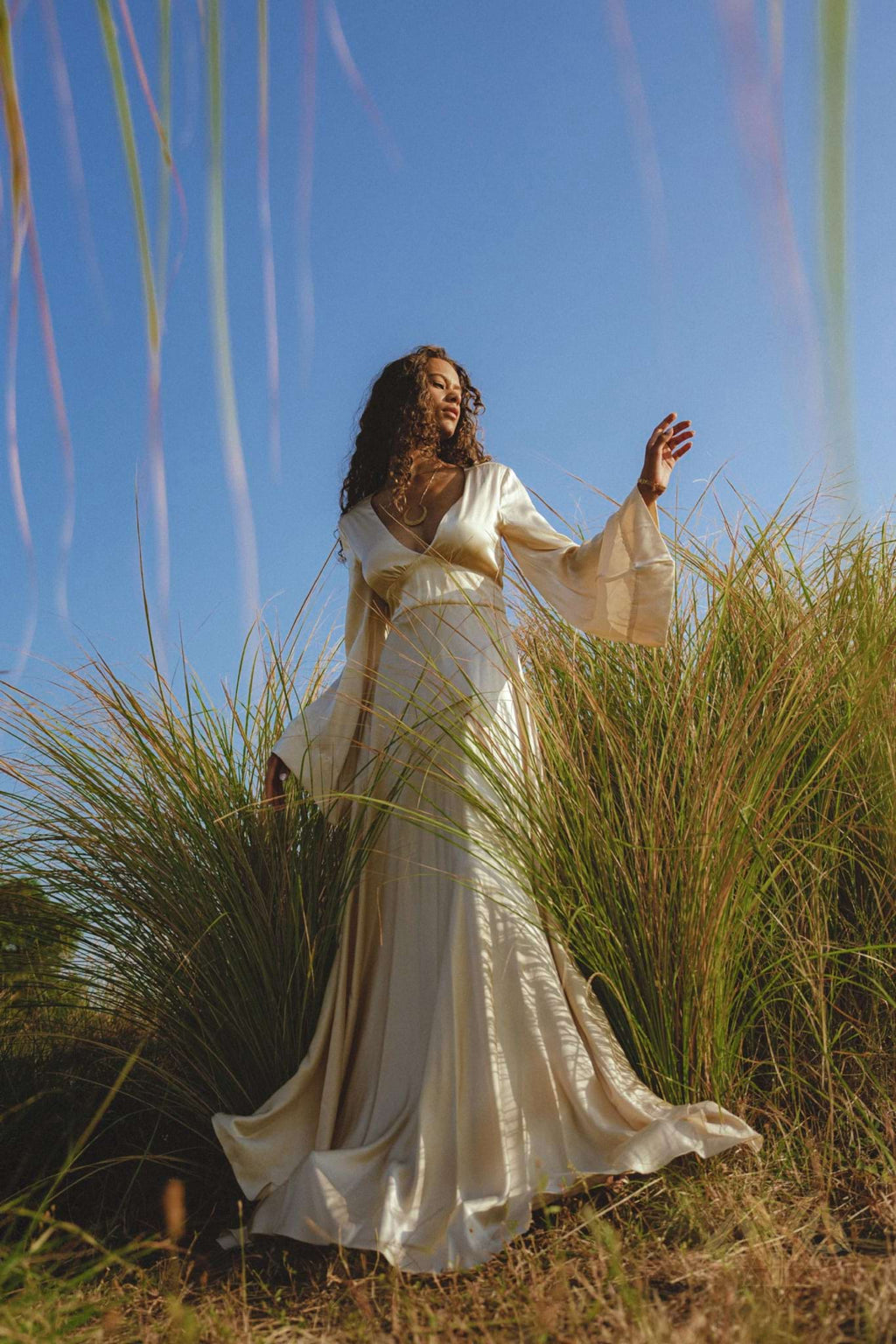 A woman in a Vanilla Cream Sundari Silk Gown by AYA Sacred Wear stands amid tall grass under a clear sky, extending her arm gracefully and gazing ahead, creating an elegant and ethereal scene.