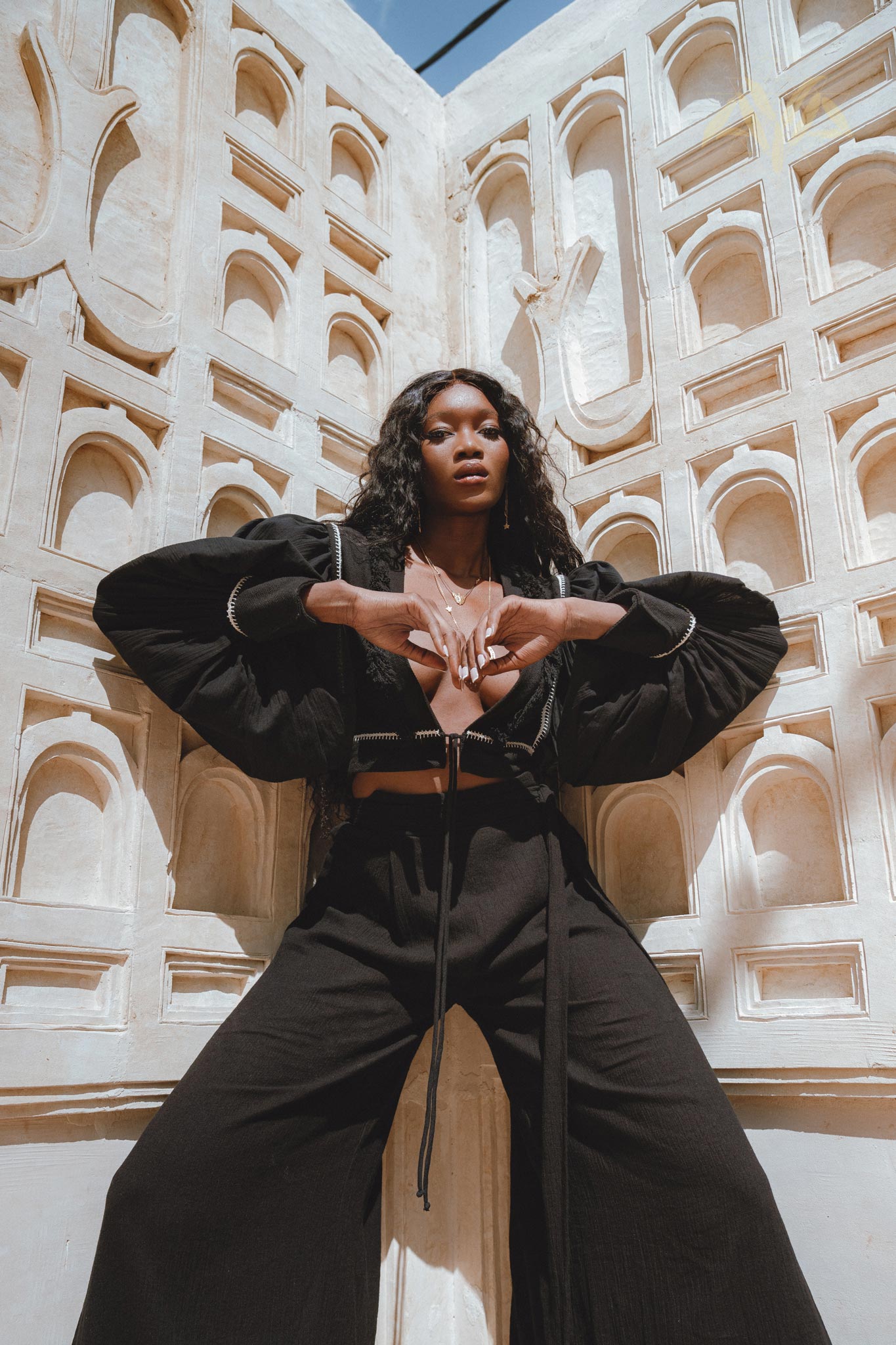 A model poses confidently in black boho pants and a matching top, showcasing a bohemian style against a textured white background. The outfit features wide-leg trousers and a cropped, layered top, emphasizing a casual yet chic look, ideal for boho fashion enthusiasts.