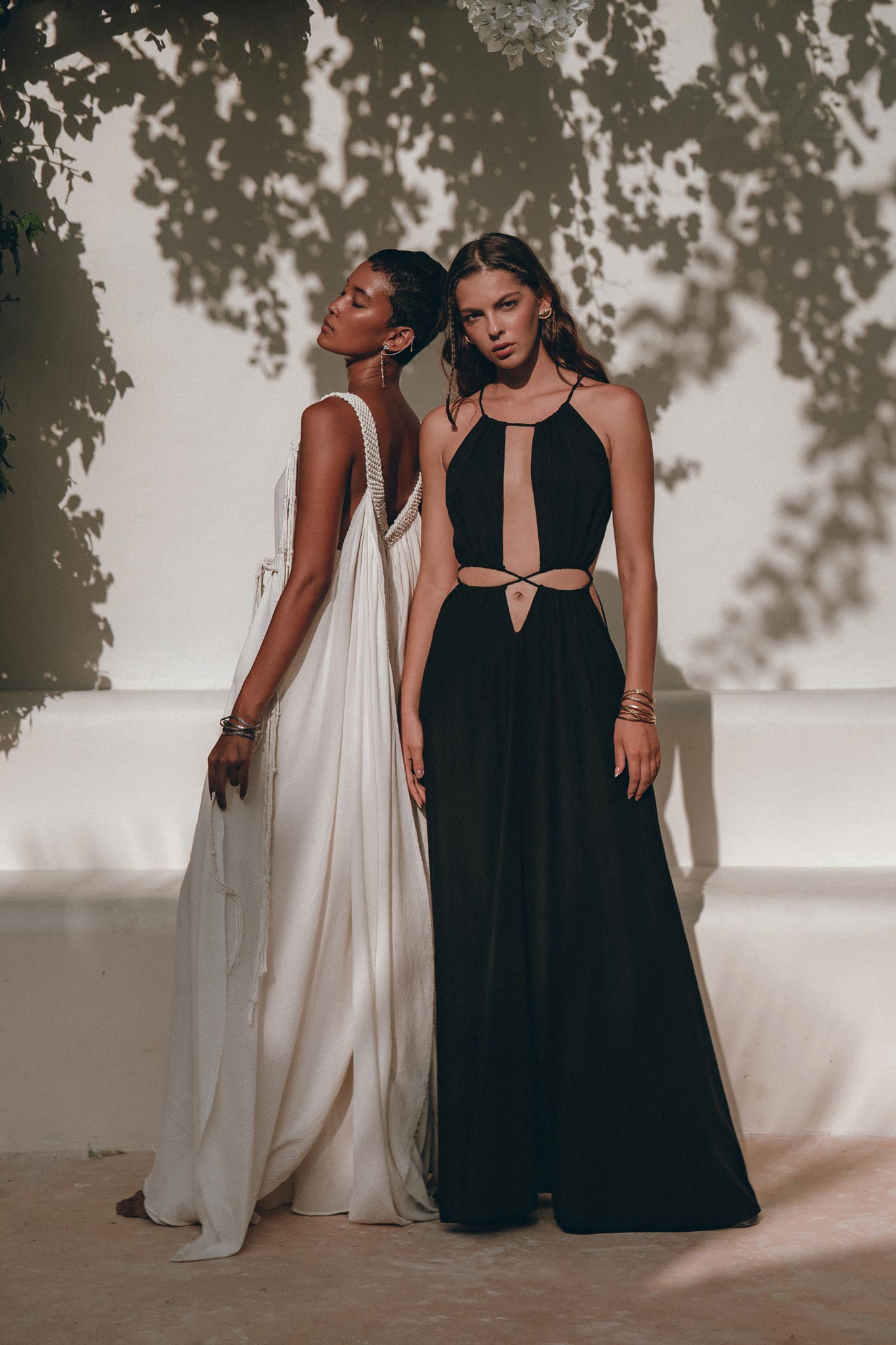 Against the textured wall outdoors, two models showcase elegant dresses made from handwoven fabrics. One model is adorned in a flowing white gown, while the other wears a stylish black prom dress with cutouts, crafted by AYA Sacred Wear and ideal for boho bridesmaids. Shadows of leaves cast a patterned backdrop on the wall.