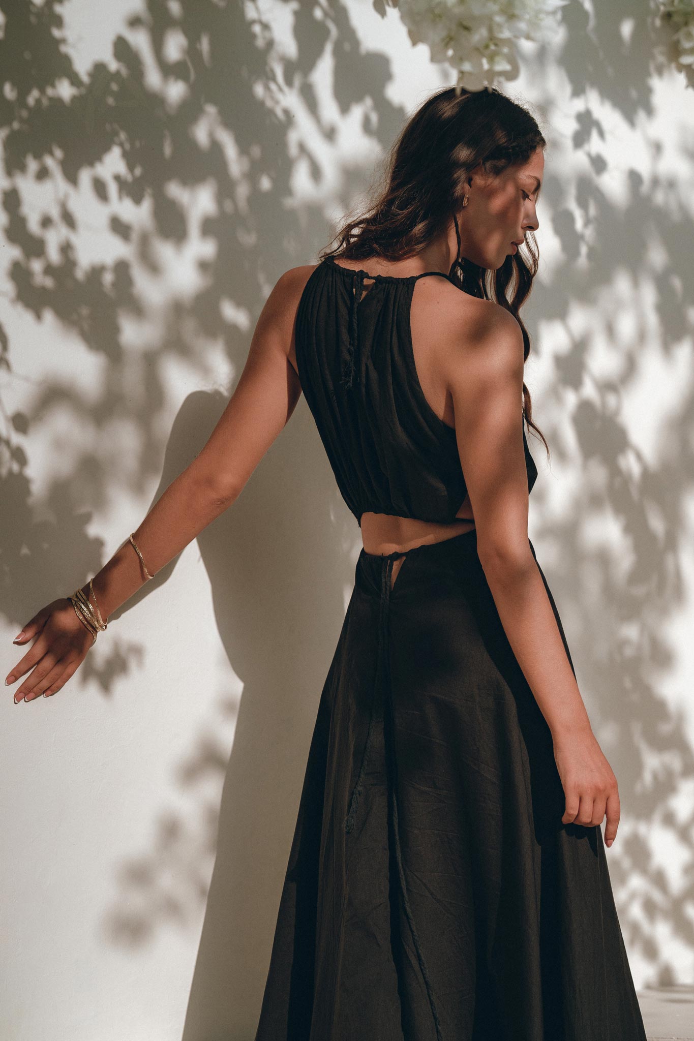 A woman wearing the Black Prom Handmade Dress from AYA Sacred Wear, a sleeveless boho dress crafted for women, poses against a white wall embellished with leafy shadows. She elegantly extends one arm, her dark hair flowing down her back in this serene and sunlit scene.