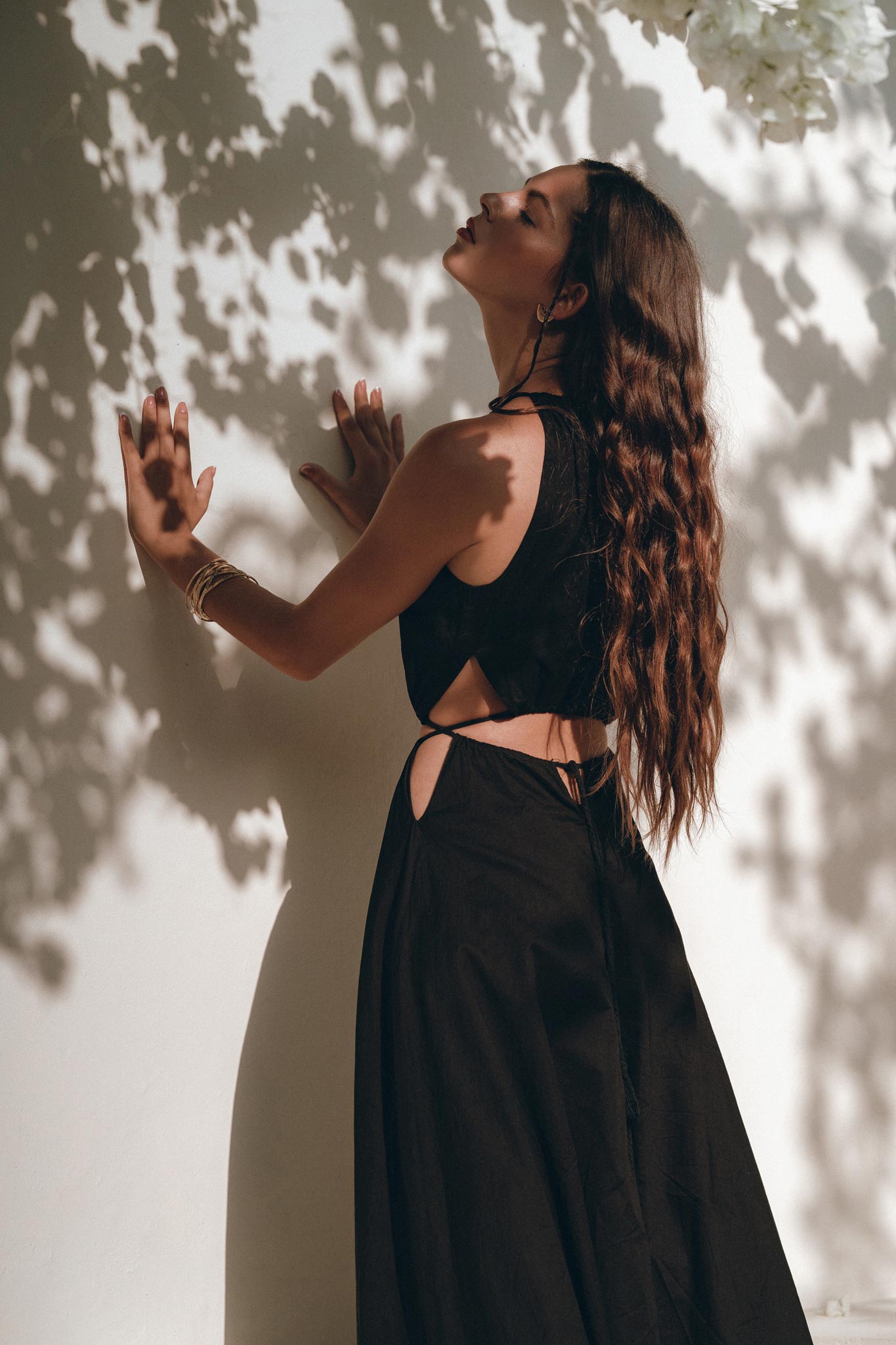 A woman wearing the Black Prom Handmade Dress by AYA Sacred Wear stands against a white wall, with dappled sunlight casting leaf patterns across its surface. Her long, wavy hair frames her relaxed expression as she gently touches the wall.