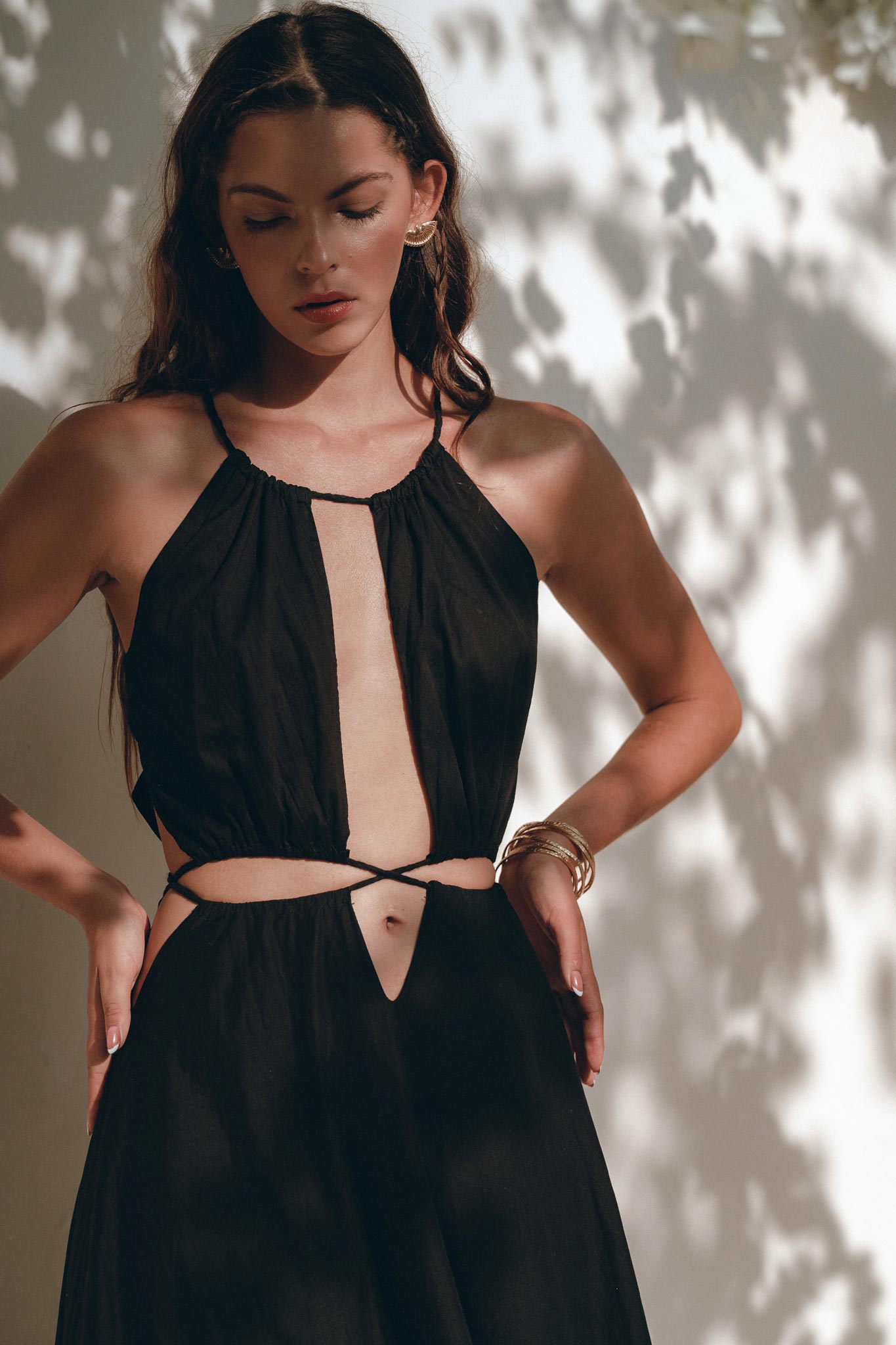 A woman in a sleeveless Black Prom Handmade Dress by AYA Sacred Wear, crafted from organic fabric, stands against a wall with dappled sunlight creating patterns around her. She has long, dark hair and wears hoop earrings, along with bracelets on one wrist. Her hands rest on her hips as she looks downward.