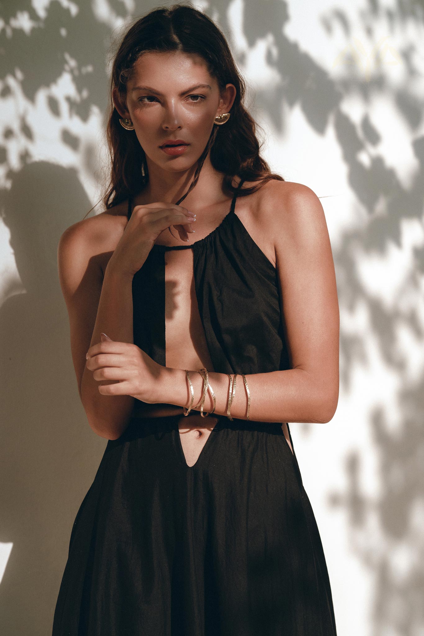 A woman wearing the Black Prom Handmade Dress by AYA Sacred Wear, crafted in a sleeveless boho style, stands against a wall partially shaded by tree leaves. Her long brown hair complements her gold hoop earrings and bracelets as she gazes directly at the camera, set against the backdrop of a warm, sunny day.