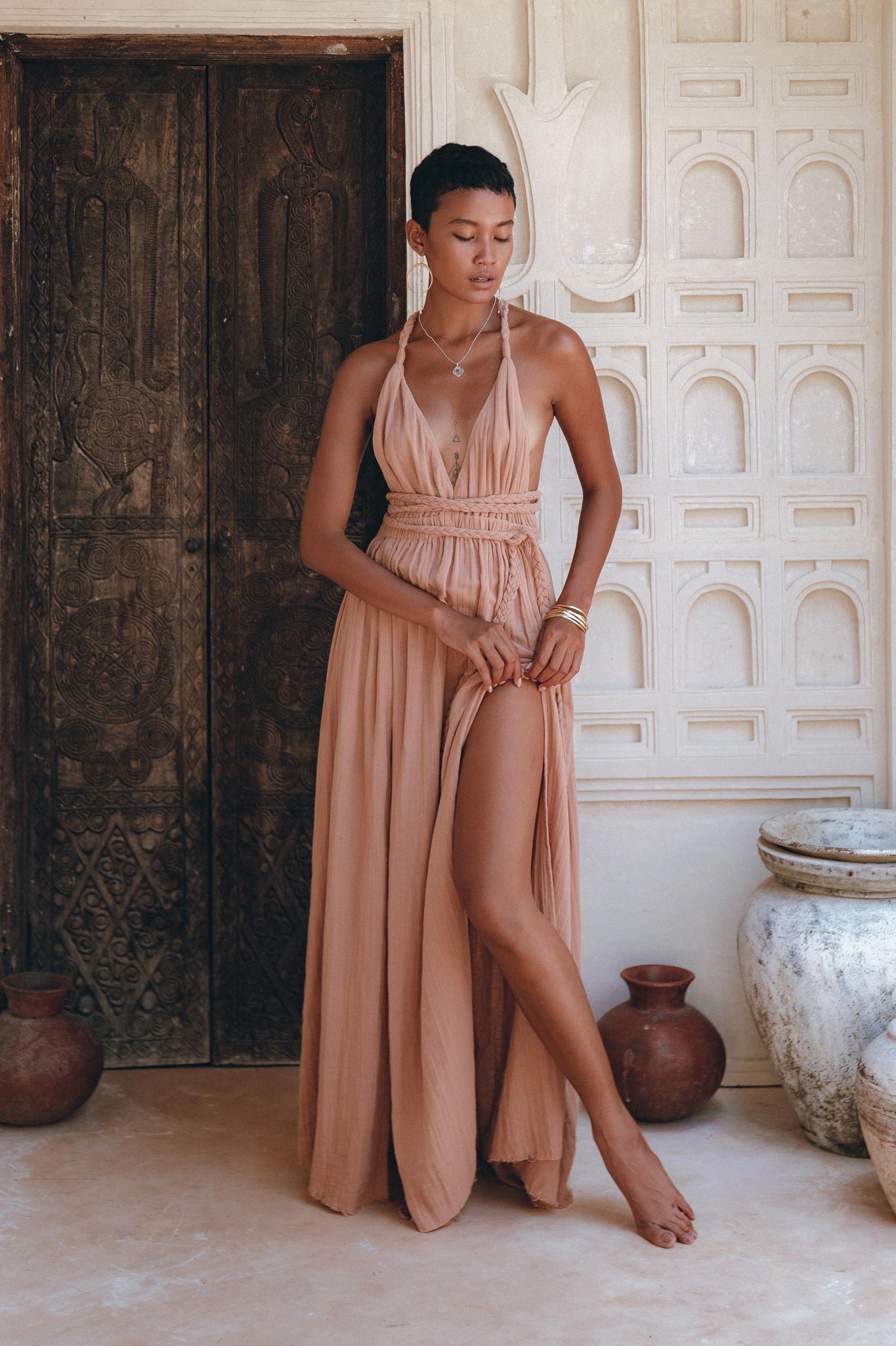 A person in a flowing Dusty Pink Greek Goddess Dress by AYA Sacred Wear stands barefoot in an elegant setting with ornate wooden doors and decorative vases. They pose gracefully with one leg slightly forward, showcasing the dress's slit. The atmosphere is serene and stylish, reminiscent of a Greek goddess's timeless charm.