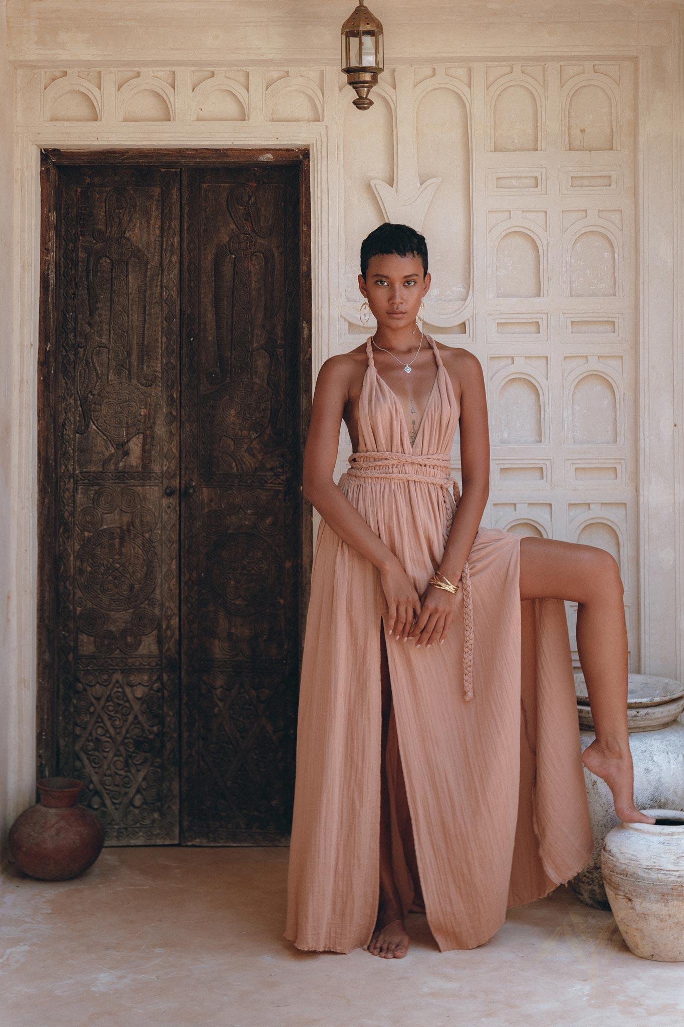 A person wearing the Dusty Pink Greek Goddess Dress by AYA Sacred Wear stands in front of an intricately carved wooden door. The background features ornate white plasterwork. With short hair and one leg raised, they elegantly showcase the dress's high slit.