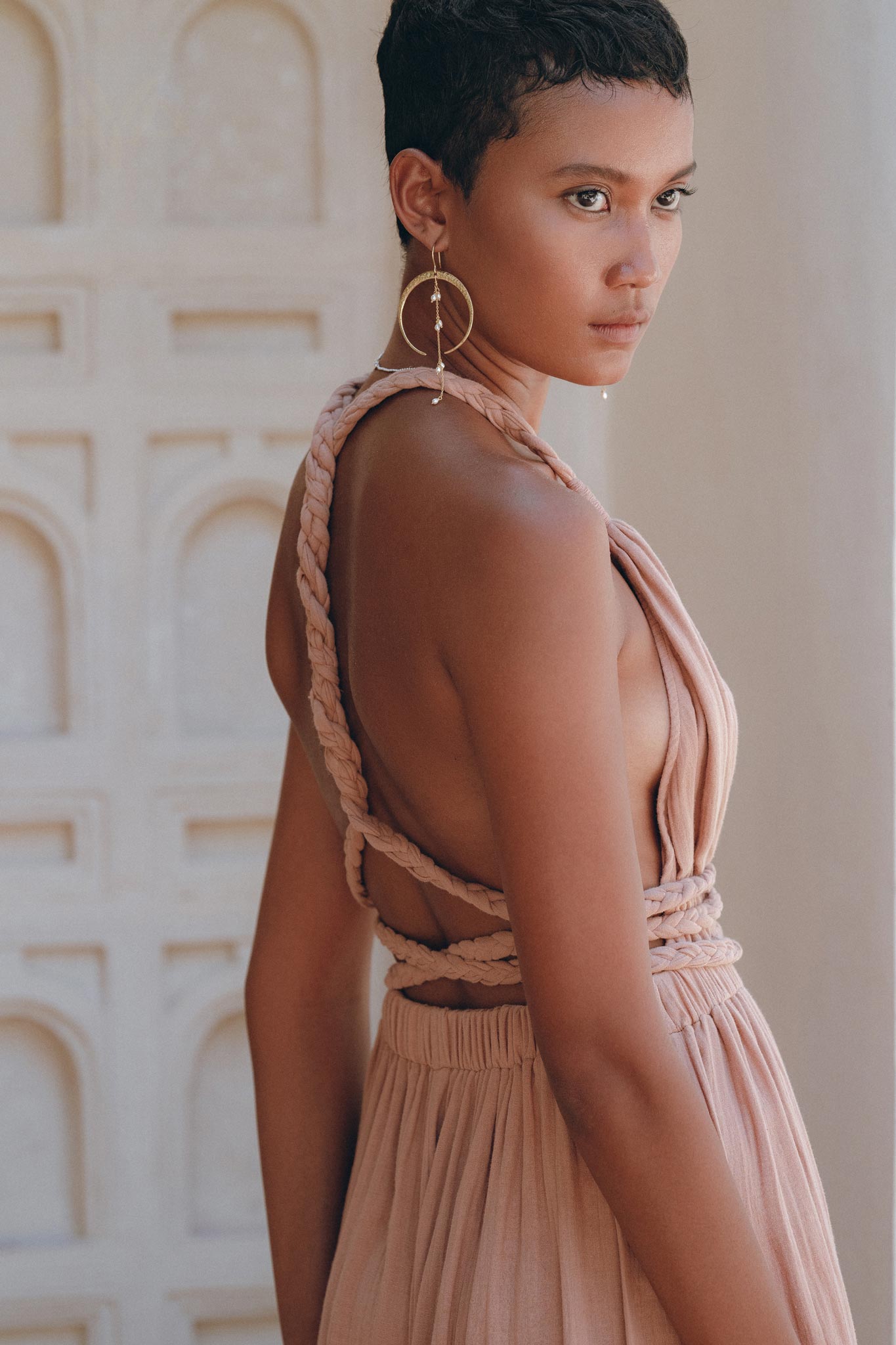 A person with short hair wearing the Dusty Pink Greek Goddess Dress by AYA Sacred Wear, styled with large hoop earrings, stands in a softly lit room looking away. The background showcases an ornate, light-colored wall adorned with arch patterns.