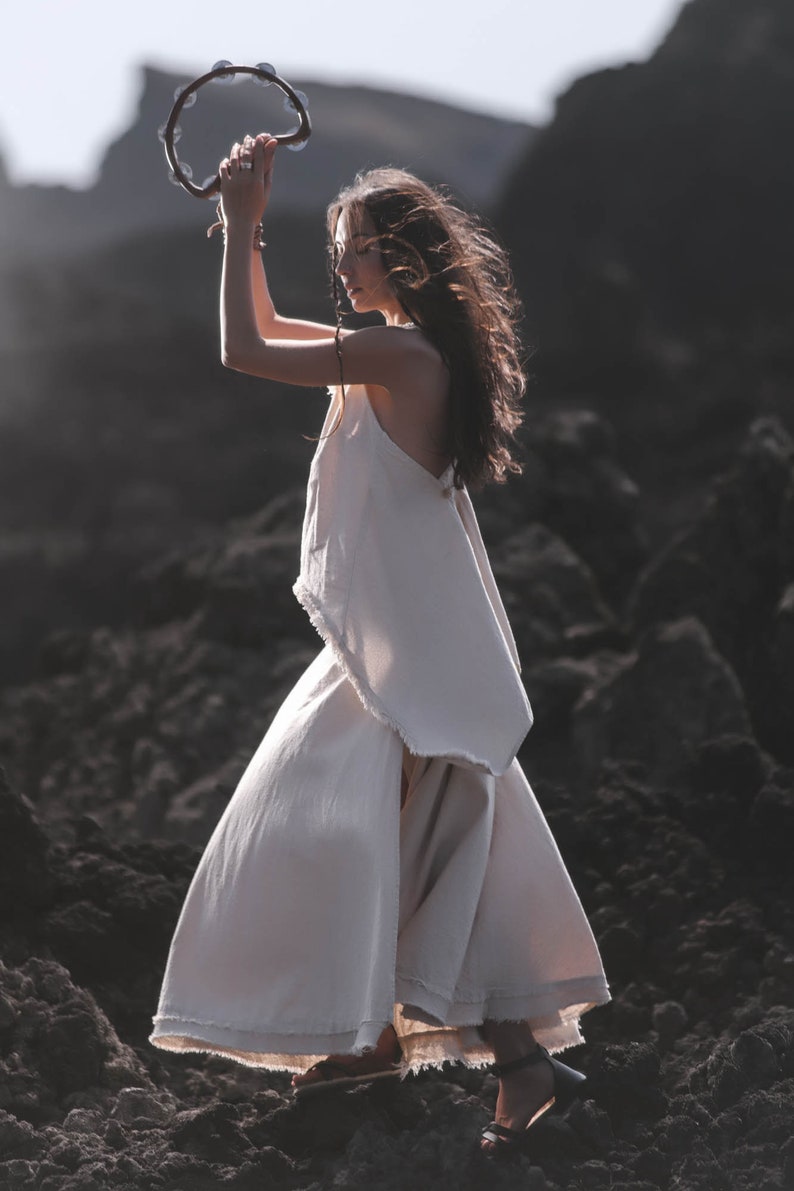 A woman wearing the AYA Sacred Wear Off-White Linen Boho Top • Viking Tunic stands on rocky terrain, holding a tambourine overhead as sunlight shines on her hair.