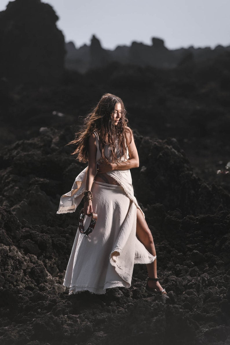 A woman in an Off-White Linen Boho Top for Women • Viking Tunic by AYA Sacred Wear stands on volcanic rocks, tambourine in hand. Soft sunlight highlights her loose hair as she gazes to the side against the dramatic landscape.