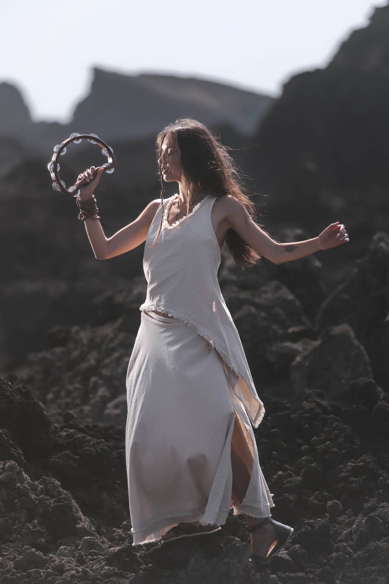 A woman stands outdoors on rocky terrain, her long hair flowing as she radiates bohemian spirit in the AYA Sacred Wear Off-White Linen Boho Top for Women • Viking Tunic, holding a tambourine under the open sky.