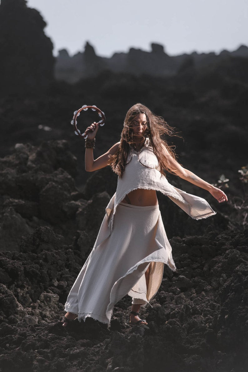 A woman wearing the AYA Sacred Wear Off-White Linen Boho Top for Women • Viking Tunic dances on dark rocky terrain, lifting a tambourine as wind flows through her hair, framed by jagged rock formations in the background.