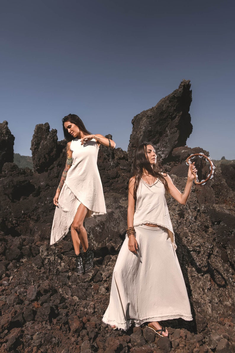 Two women in Off-White Linen Boho Tops by AYA Sacred Wear pose on rocky terrain—one with a tambourine, the other with a raised leg—showcasing Viking Tunic style against jagged rocks and a clear blue sky.