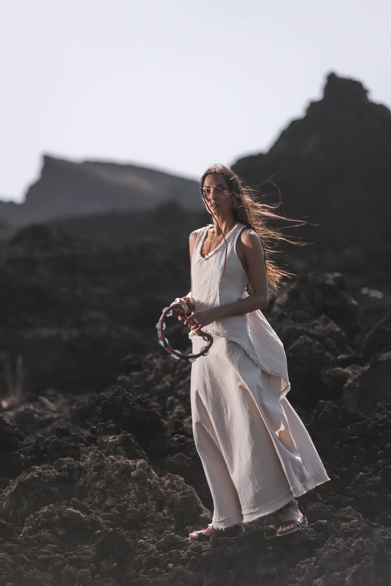 A woman wears the Off-White Linen Boho Top for Women • Viking Tunic by AYA Sacred Wear, standing on dark rocks with a tambourine as her hair blows in the wind against jagged silhouettes and a bright sky.