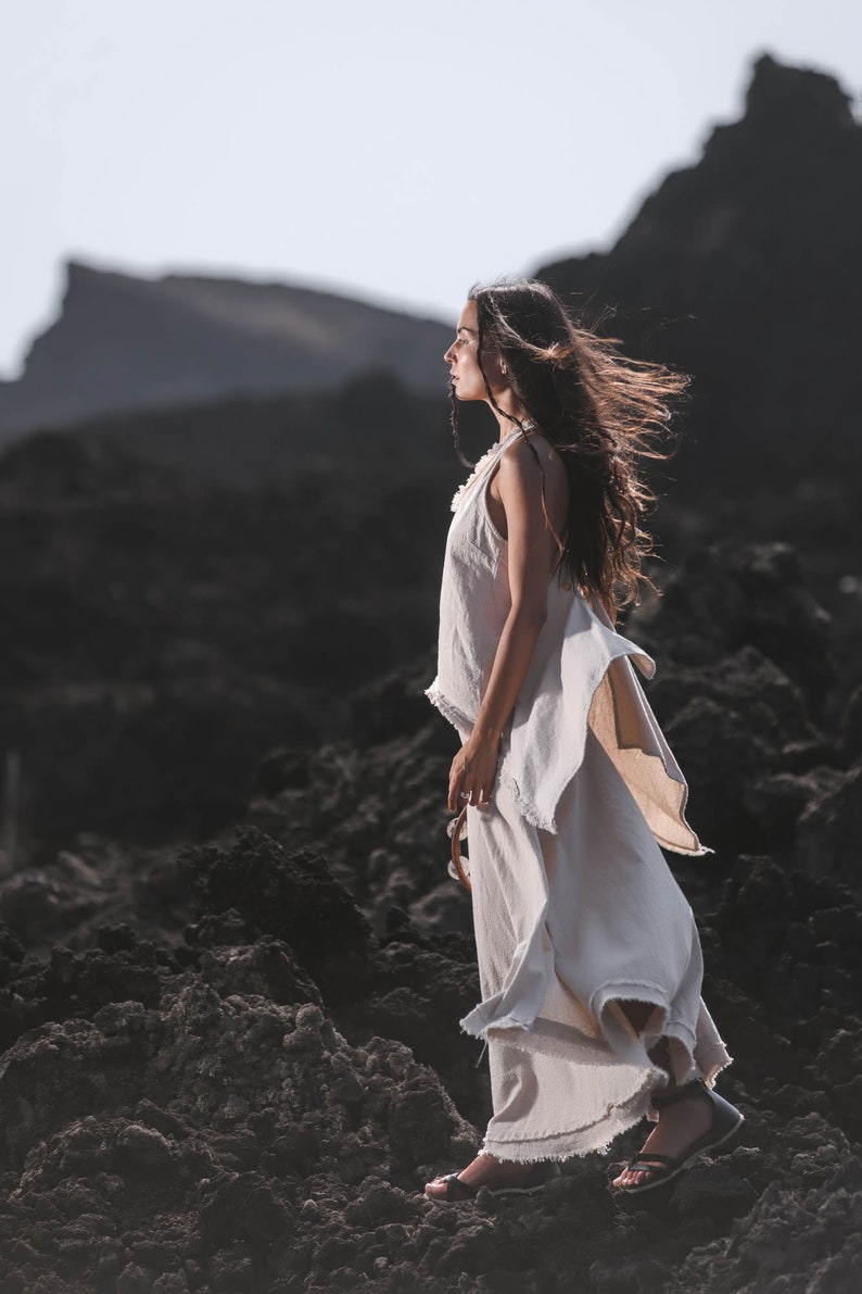 A woman wearing the Off-White Linen Boho Top for Women • Viking Tunic by AYA Sacred Wear stands on dark, rocky terrain with windswept hair and rugged hills under a bright sky.