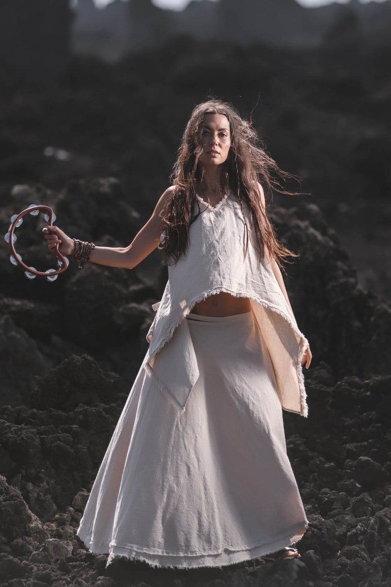 A woman with long hair stands outdoors on rocky terrain, wearing the Off-White Linen Boho Top for Women • Viking Tunic by AYA Sacred Wear, holding a tambourine and gazing confidently at the camera.