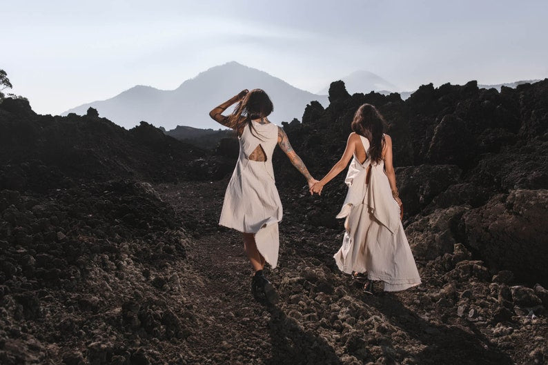 Two women wearing AYA Sacred Wear Off-White Linen Boho Tops and Viking Tunics walk hand in hand along a rocky path, surrounded by dark volcanic rocks, with mountains and a hazy sky in the background.