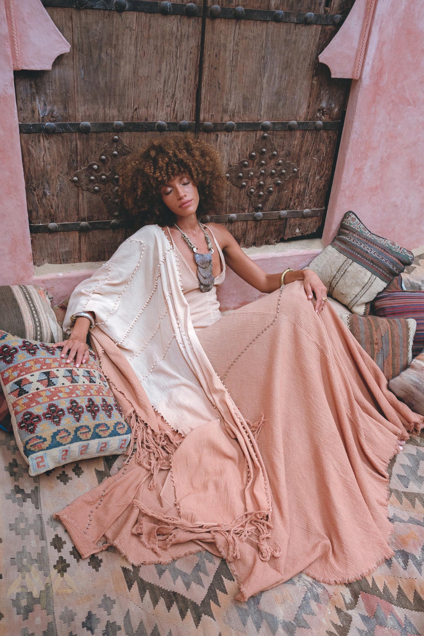 A woman with curly hair relaxes on patterned cushions in a flowy Powder Pink Boho Poncho by AYA Sacred Wear, crafted sustainably with ombre botanical dyes. She's enveloped by rustic wooden doors and textured textiles, creating a bohemian ambiance.