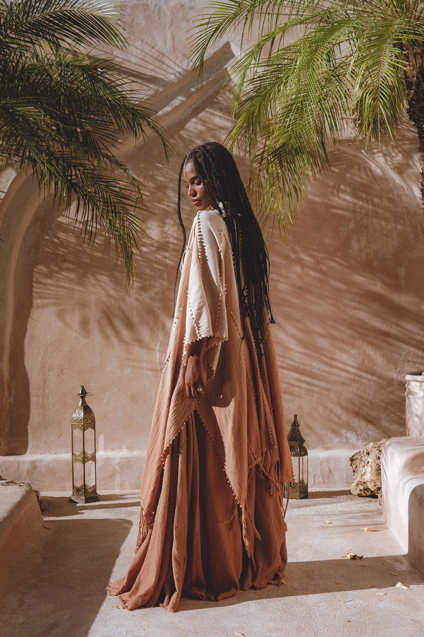 A person with long braided hair stands outside in a sunlit courtyard, wearing an AYA Sacred Wear Powder Pink Boho Poncho crafted from botanical dyeing techniques. They are surrounded by lush palm plants and decorative lanterns that cast soft shadows on the pale wall.