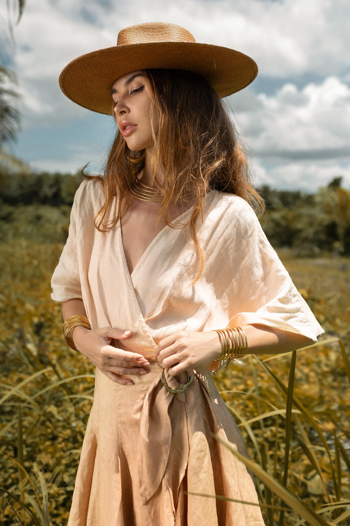 A woman stands in a field under a clear sky, wearing the Ombre Light Pink Dyed Mini Wrap Dress from AYA Sacred Wear. She has long hair and is accessorized with multiple bangles and a wide-brimmed hat. She appears serene, with her hands gently resting on her midsection.